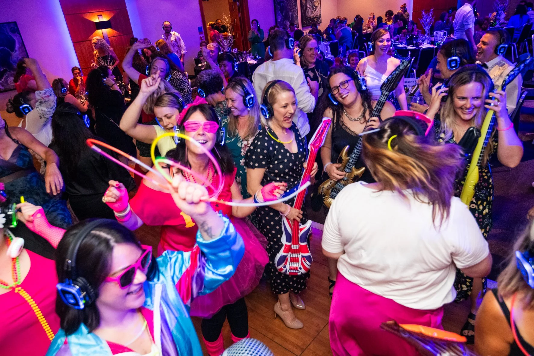 People enjoying a lively dance party, wearing colorful costumes, sunglasses, and headphones, with music instruments like guitars, dancing, and happy expressions in a vibrant, crowded event space.