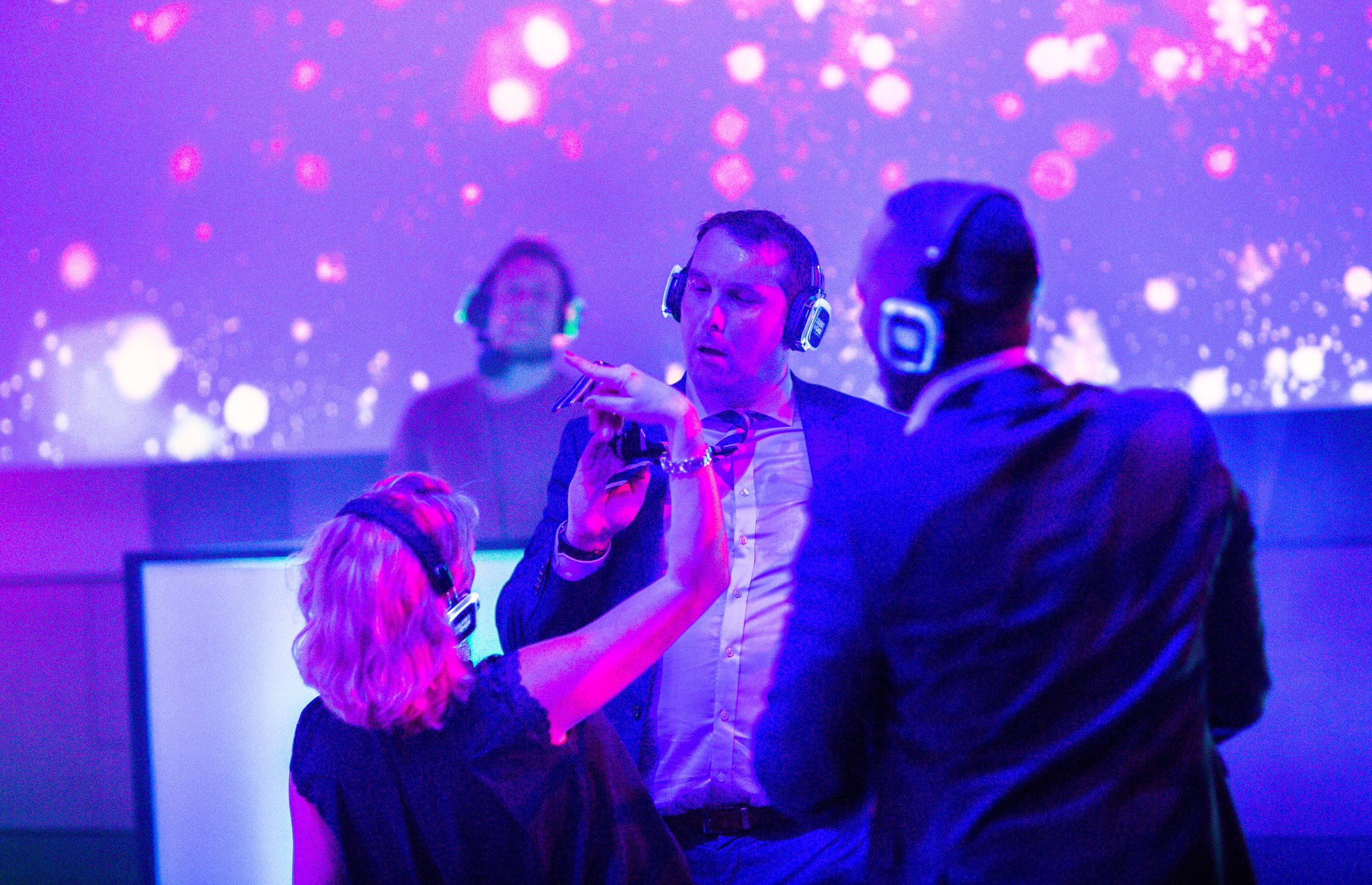 Three people wearing headphones are dancing and engaging with each other in a colorful, vibrant setting with a purple and pink background, excited and enjoying the moment.