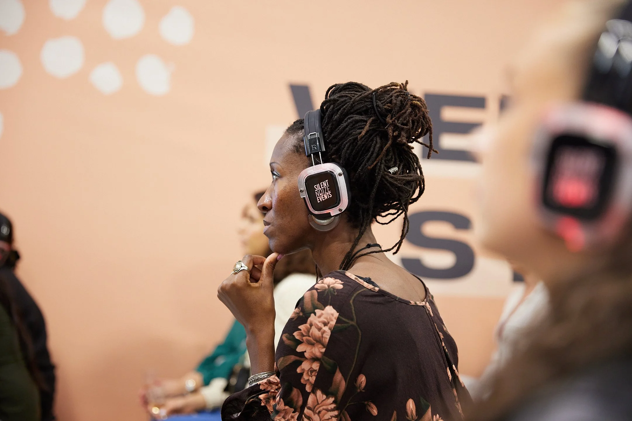 A woman with long dreadlocks wears headphones with a label that reads 'Silent Noize Events' during an indoor event, standing in profile with a floral patterned top.