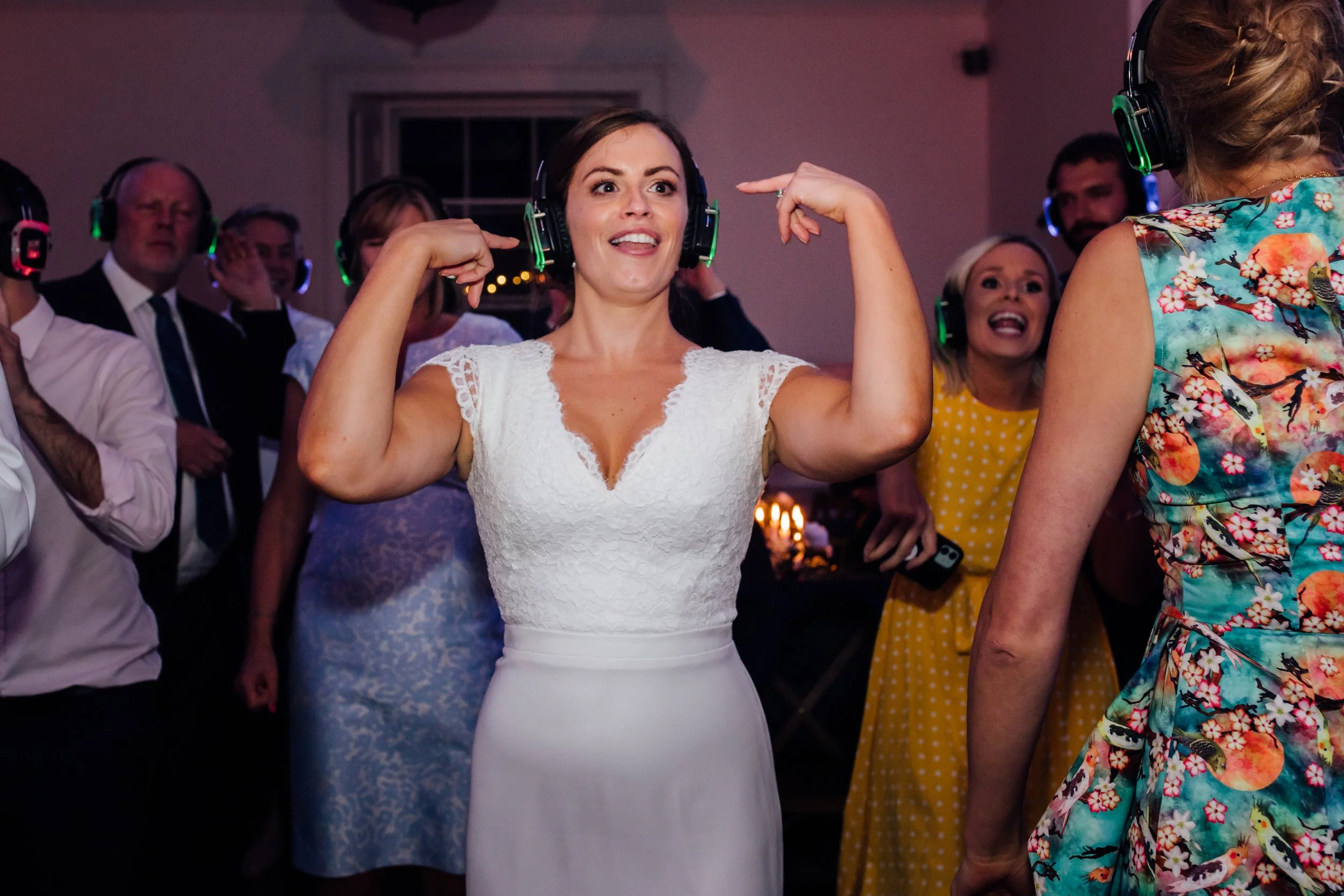 A woman in a white dress dancing at a party, wearing large headphones, with a group of people dancing and smiling behind her.