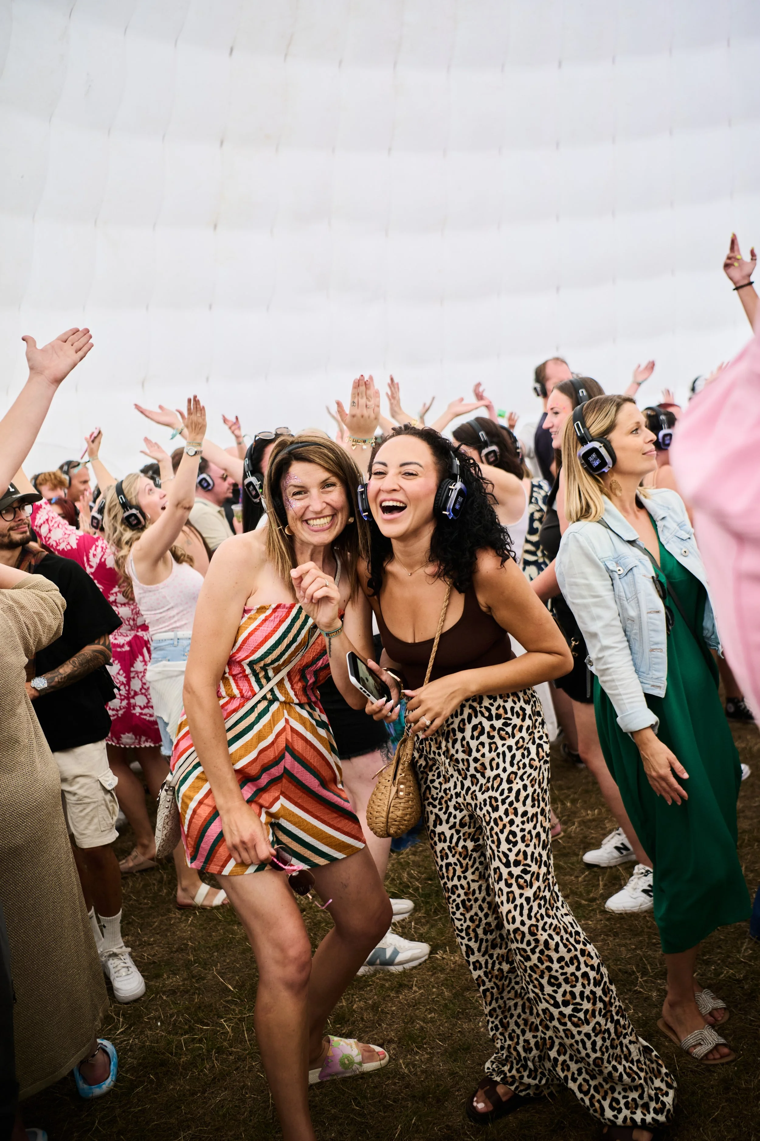 People dancing and enjoying music at an outdoor event, wearing headphones, with two women in the foreground smiling and laughing.