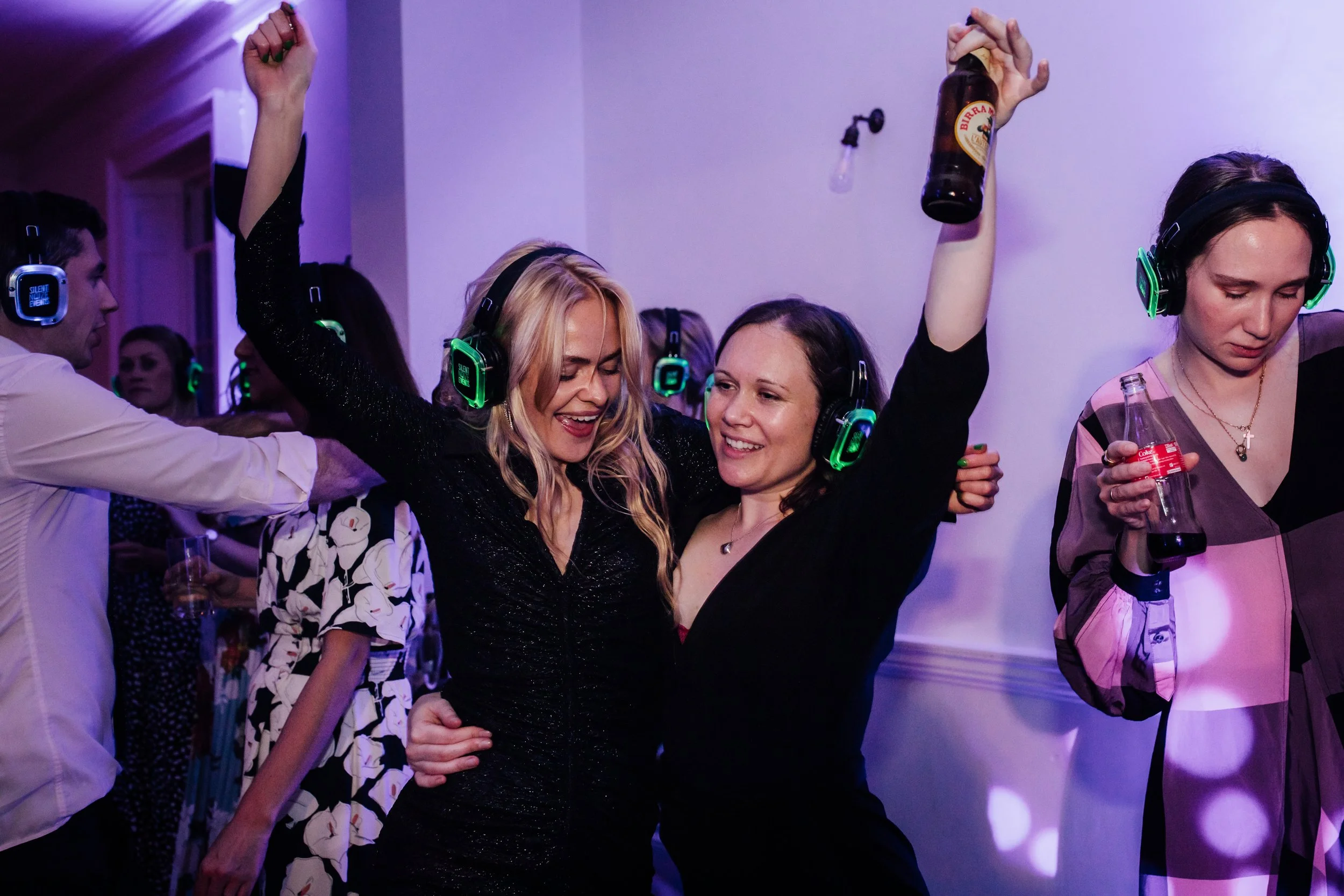 Two women dancing and smiling, wearing headphones with green lights, at a party or nightclub. One woman is holding a beer bottle, the other has her arm raised.