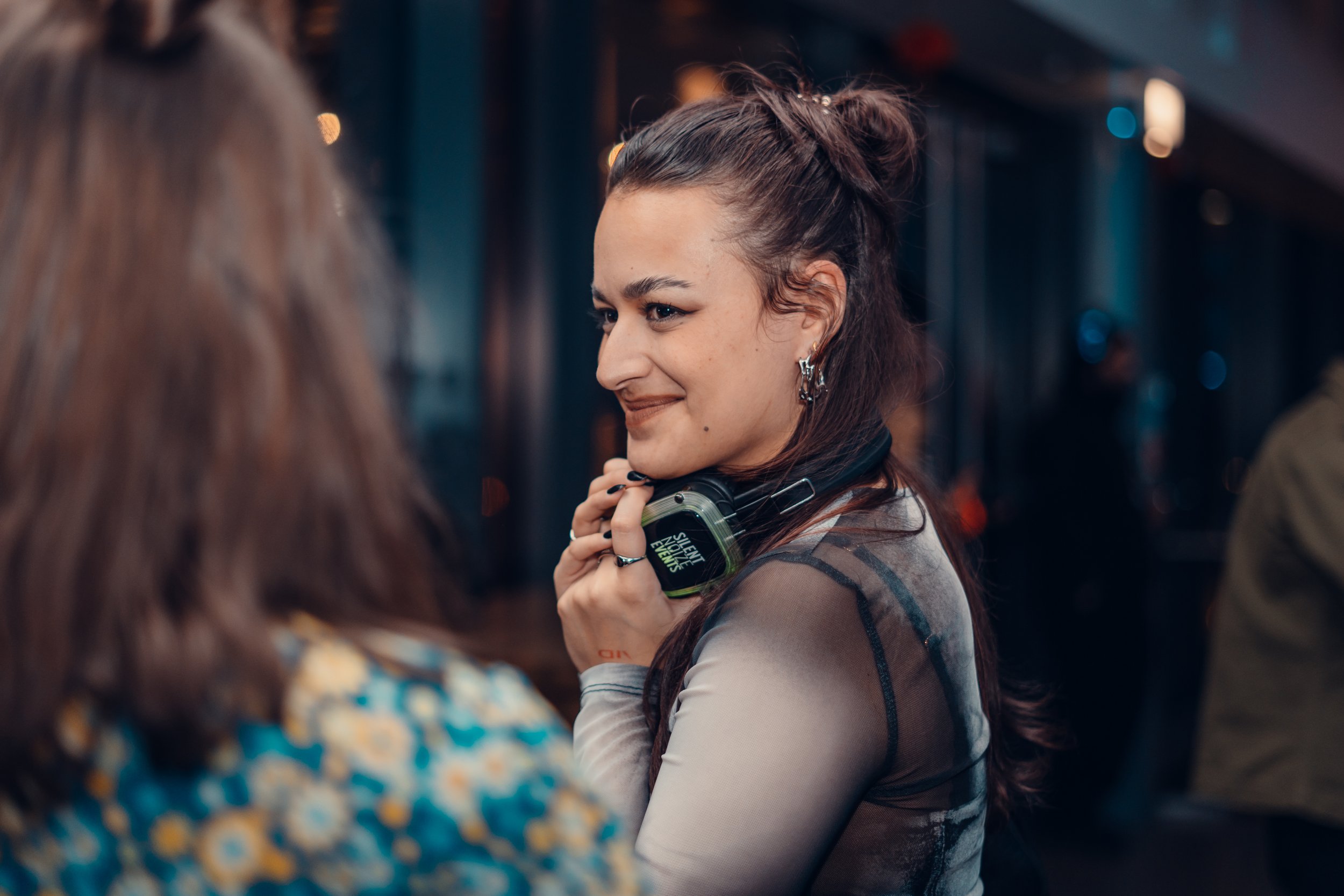 A woman with dark hair tied in a bun, earrings, and a sheer top, holding headphones around her neck, smiling and looking at another woman in a setting with dim lighting and blurred background.