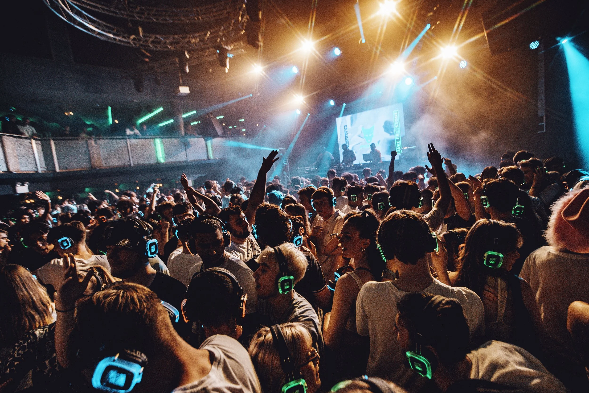 Crowd of people wearing headphones at a concert or music event, with stage lights and a large screen in the background.