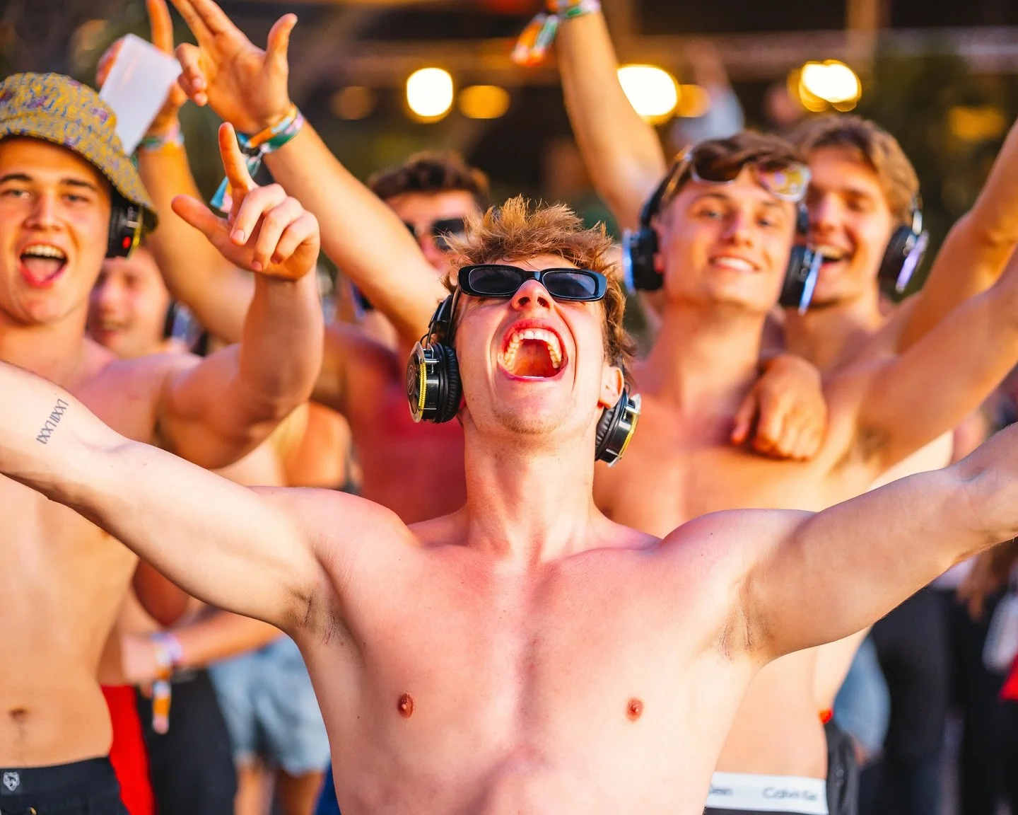 A group of young men dancing and celebrating outdoors at a music festival, with some wearing headphones and sunglasses, shirtless with arms raised, smiling and enjoying the moment.
