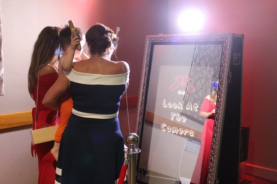 People looking at a mirror with a sign that says 'Look at the Camera' in a decorated frame.