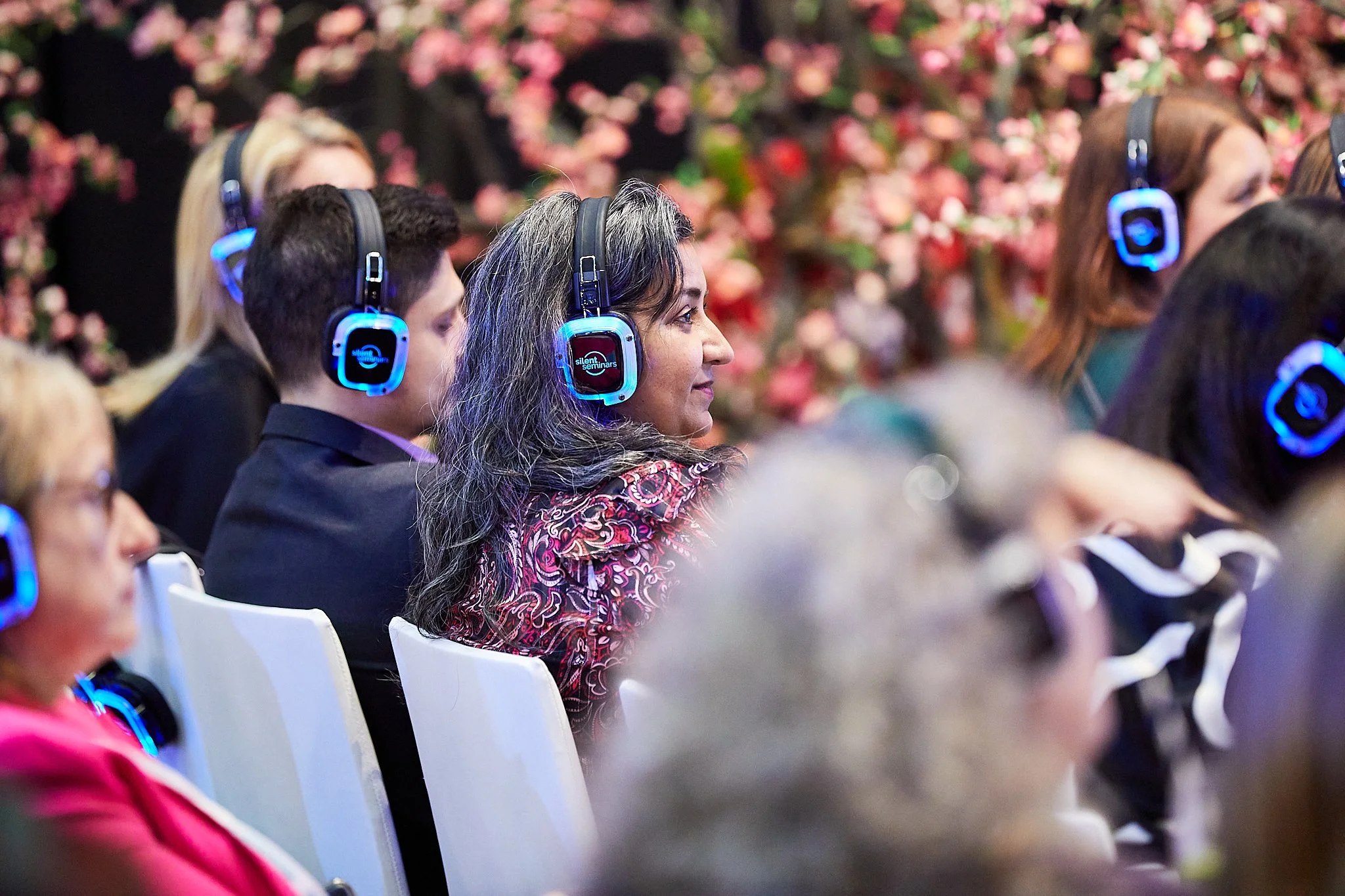 People seated in an audience attending a presentation or conference, wearing wireless headphones, with a background of pink flowers.