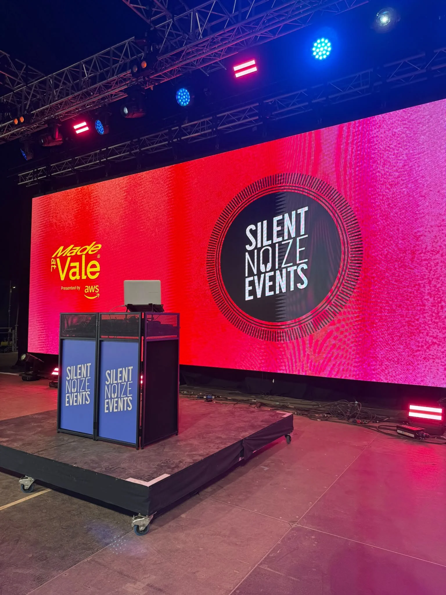 Stage setup with a large LED screen displaying the words 'Silent Noize Events' in the center and 'Made at Vale' promoted by AWS on the left. There are two smaller screens in front with the same text. Stage equipment and cables are visible on the floor.