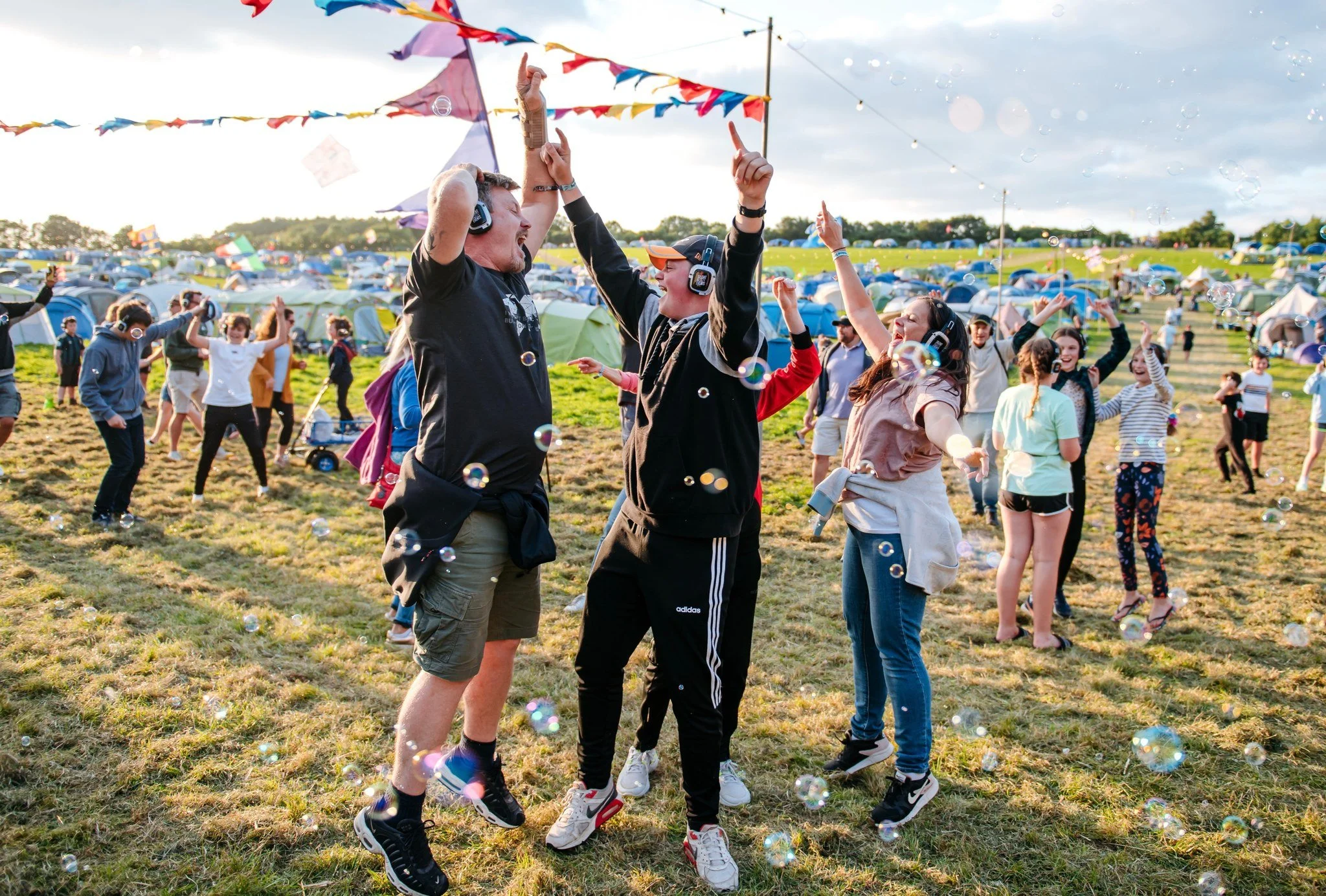 People dancing and celebrating at an outdoor festival with tents and colorful banners in the background, under a partly cloudy sky, with bubbles floating around.