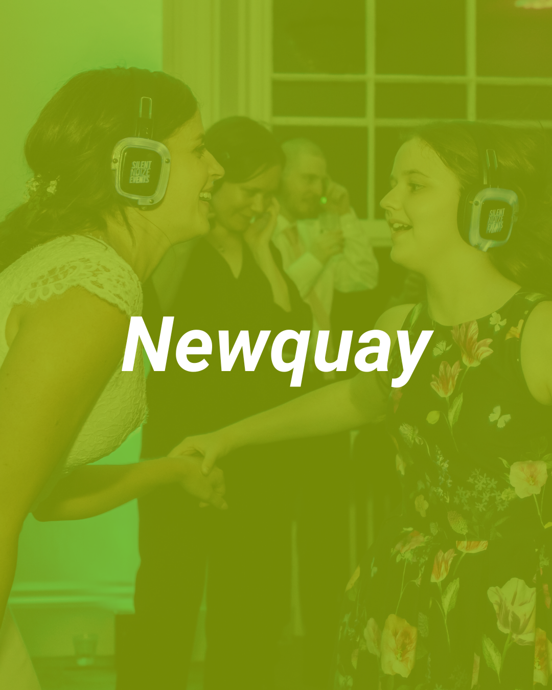 Two women wearing headphones are smiling and talking to each other at a social gathering. Several people are in the background, some taking photos.