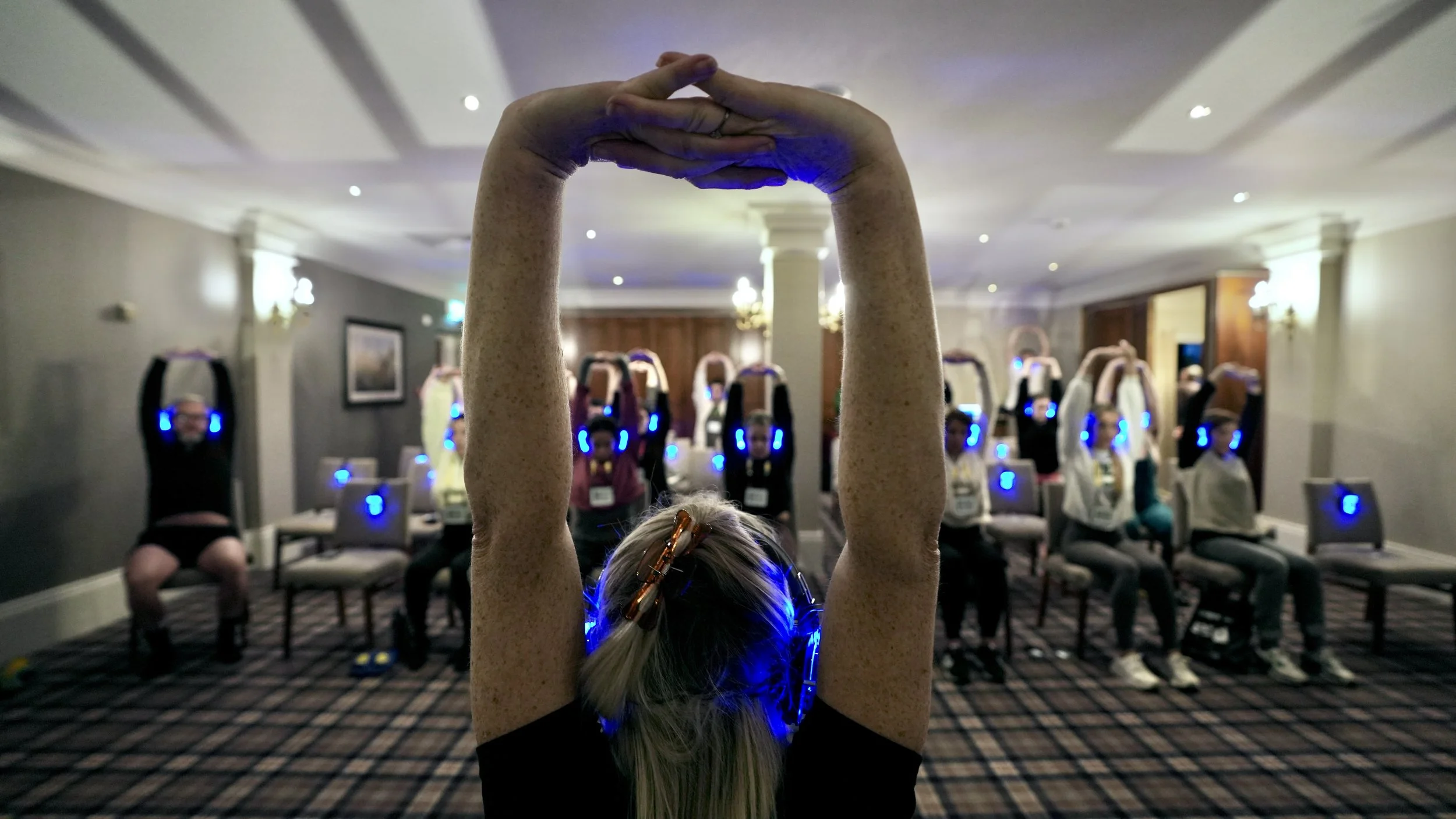People participating in a seated yoga or meditation session while wearing blue LED headphones in a conference room.