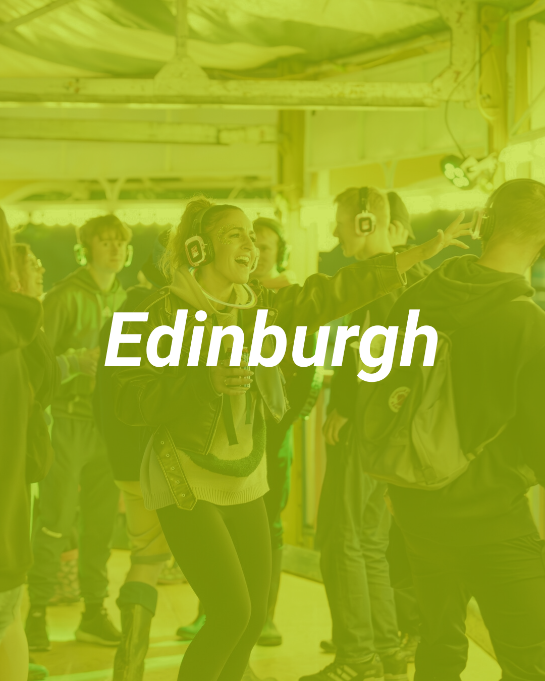 A group of young people, wearing headphones and casual clothing, engaging in an indoor activity at an event in Edinburgh. One woman appears to be leading or instructing, with her arm outstretched and a cheerful expression.