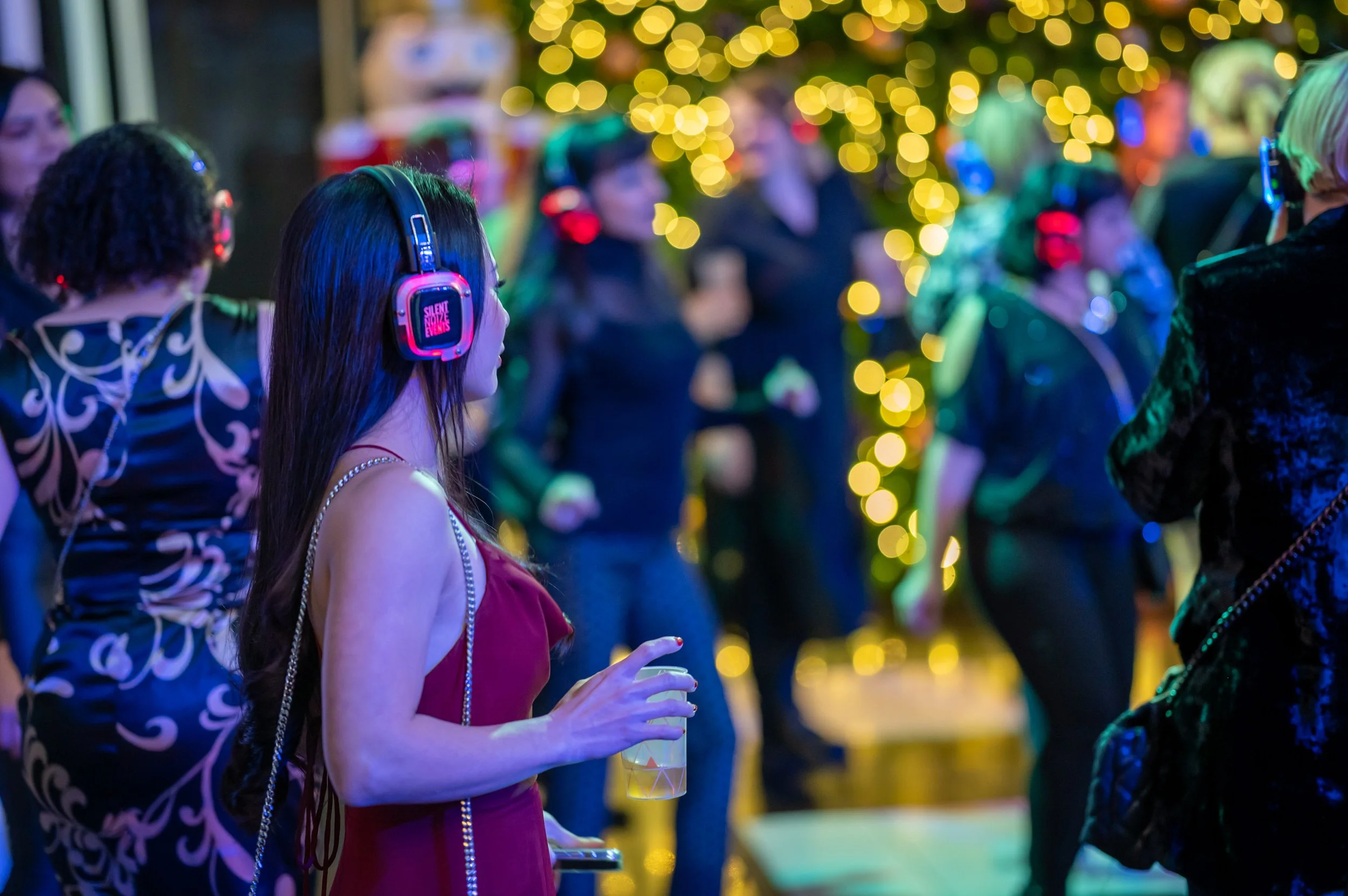 A woman with long dark hair, wearing a red dress and pearl necklace, is holding a drink while wearing headphones with pink lights. She is in a crowded setting with blurred background lights and people.