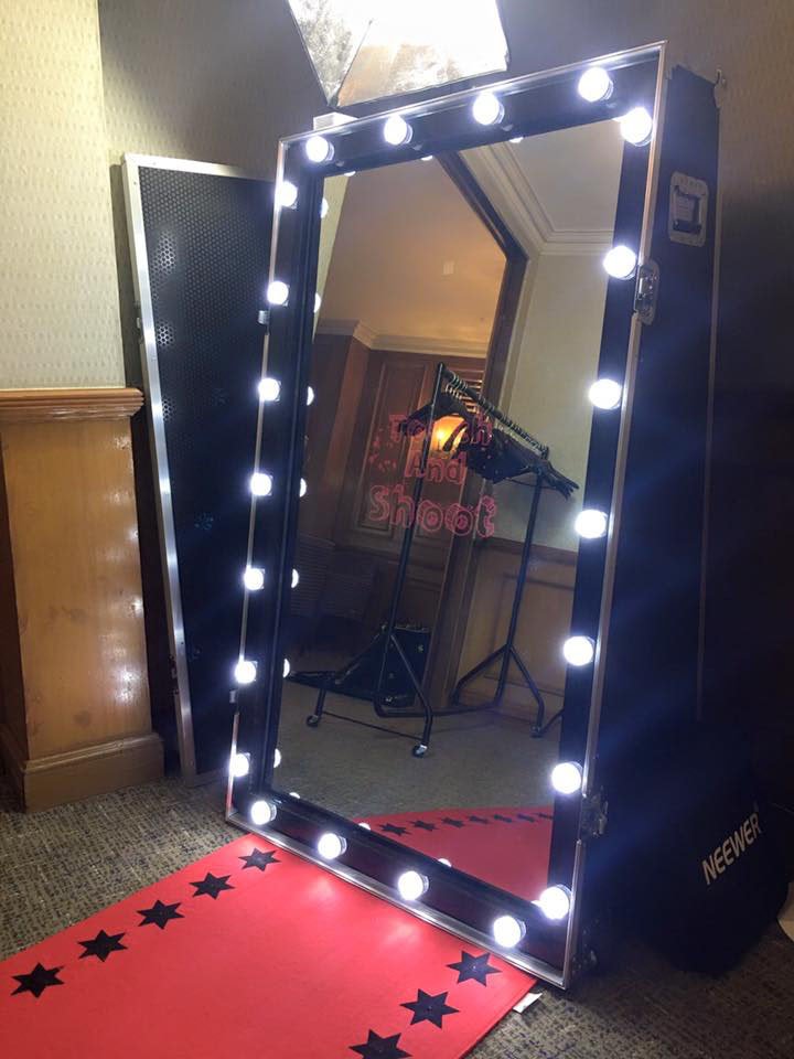 A large mirror with bright light bulbs around the frame, placed on a red carpet with black stars, in an indoor setting with a wooden wall and equipment reflected in it.