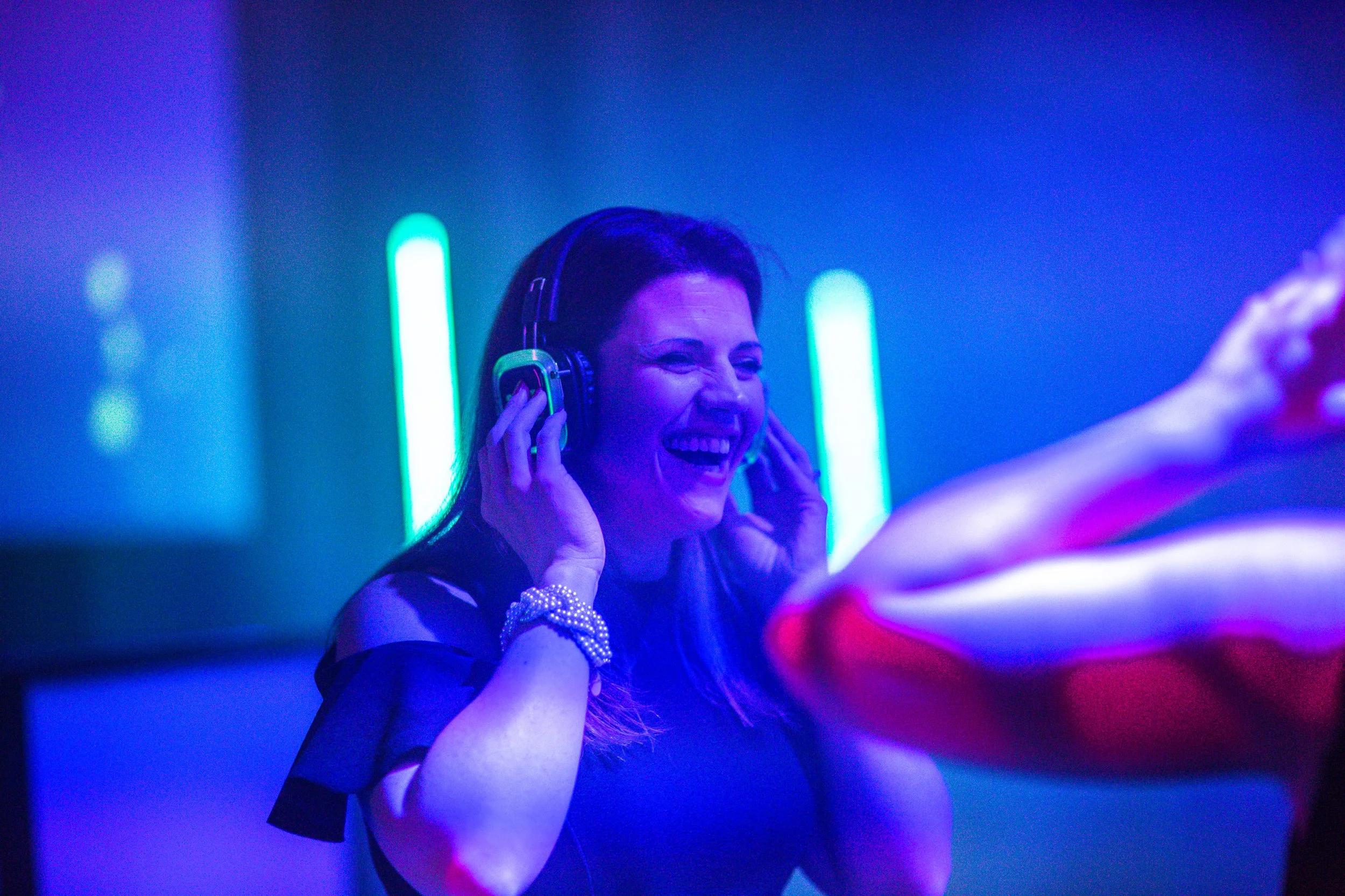 A woman with brown hair is wearing headphones and smiling at a club or concert with neon blue and green lighting.