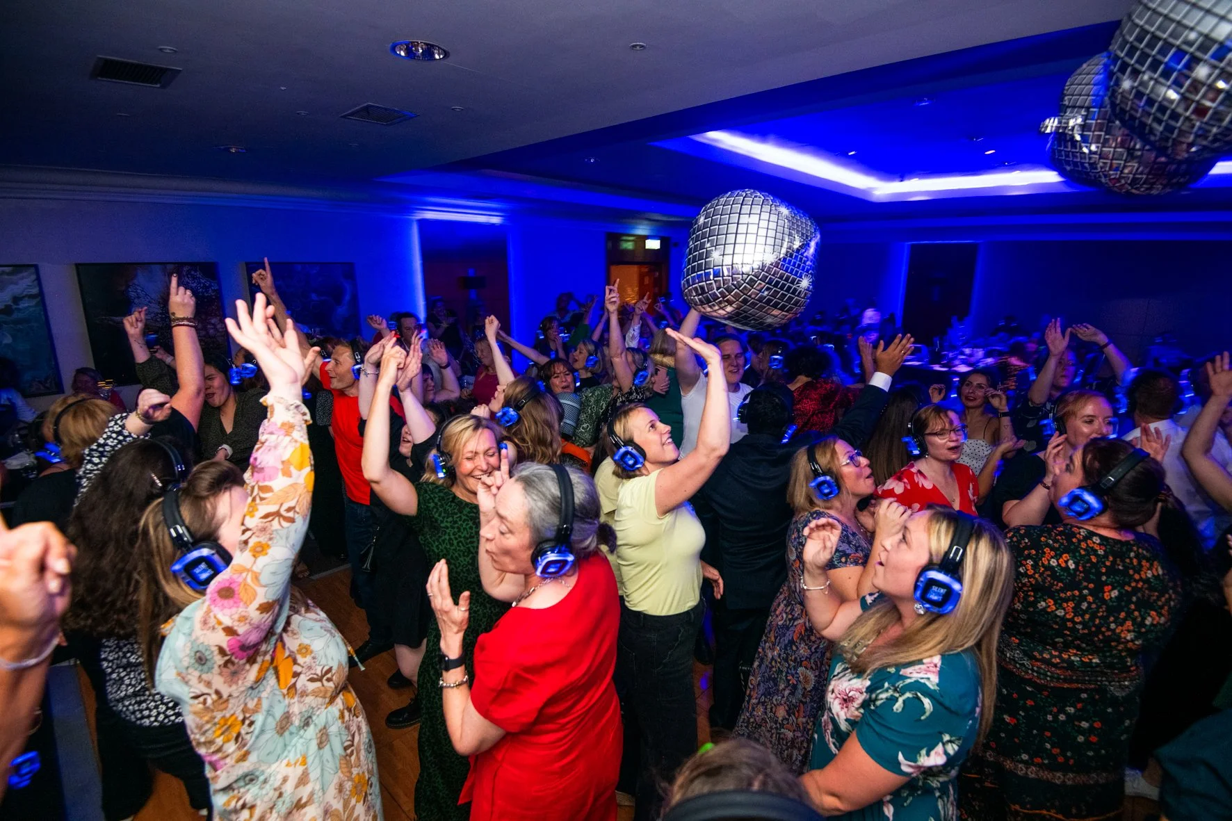 Crowd dancing and celebrating at a party with disco balls and blue lighting, many wearing headphones.