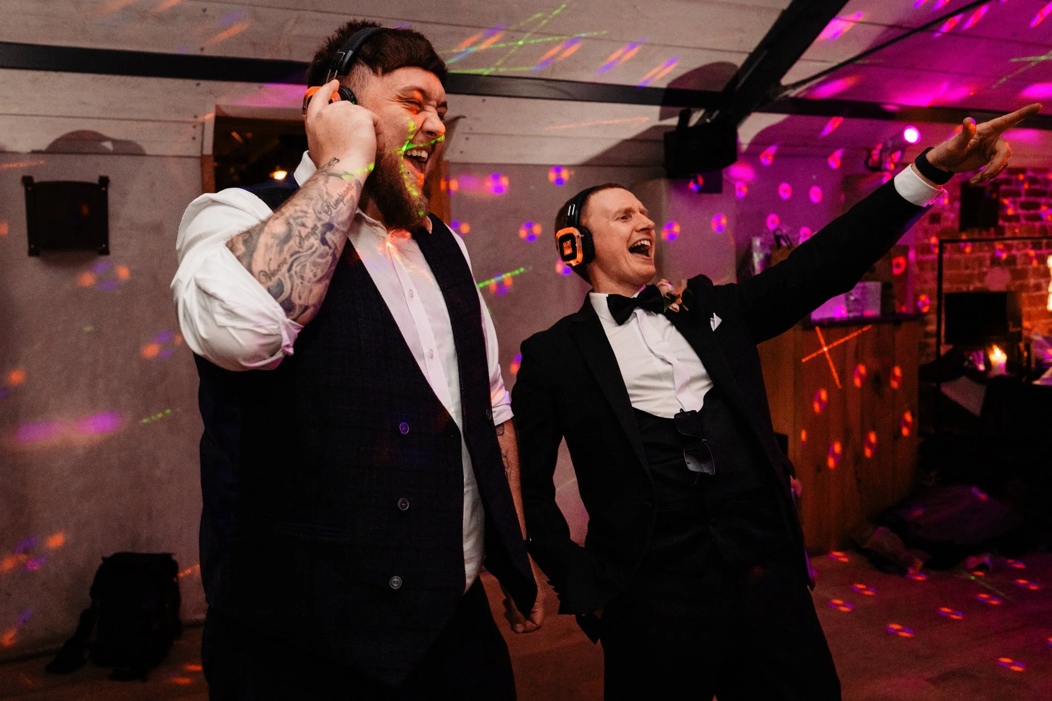 Two men in formal attire dancing and enjoying themselves at a party or celebration, with one wearing headphones and colorful laser lights in the background.