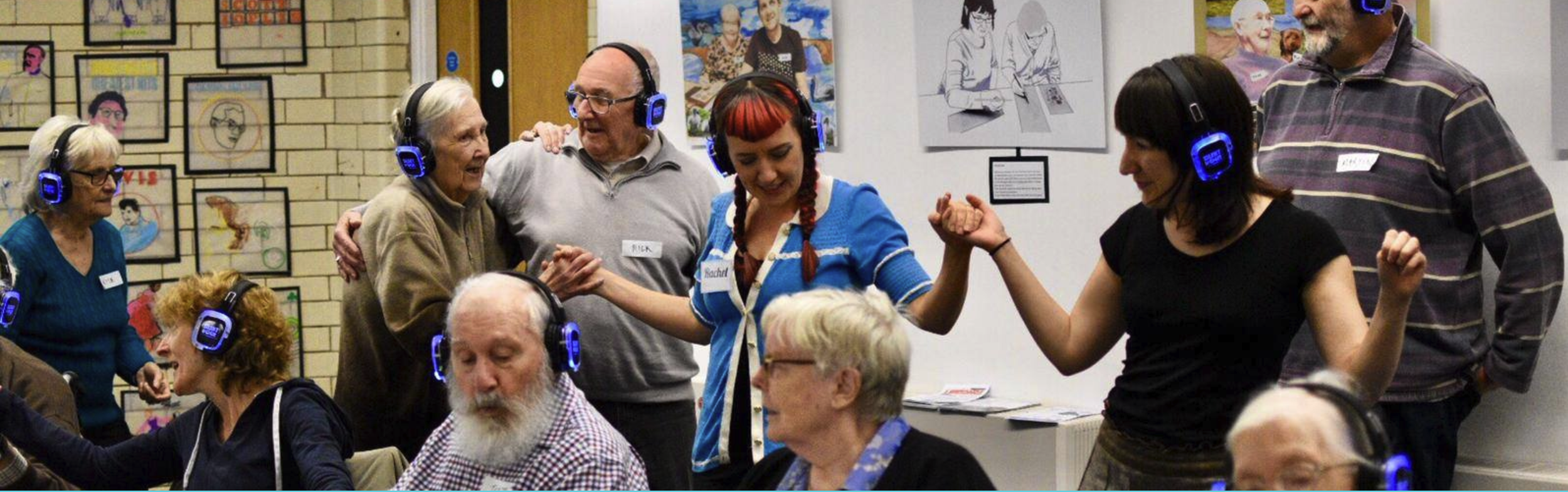 Group of elderly and young adults wearing blue headphones at a social gathering, dancing and chatting with one another, in an art exhibition space.