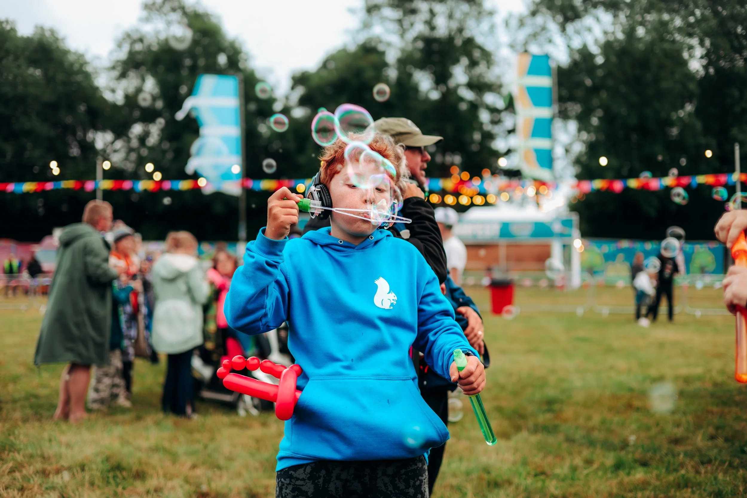 A young boy in a blue hoodie is blowing bubbles at a fair or festival, with a balloon sword hanging from his waist. In the background, there are people, colorful banners, and a blue and yellow tent, indicating an outdoor event.