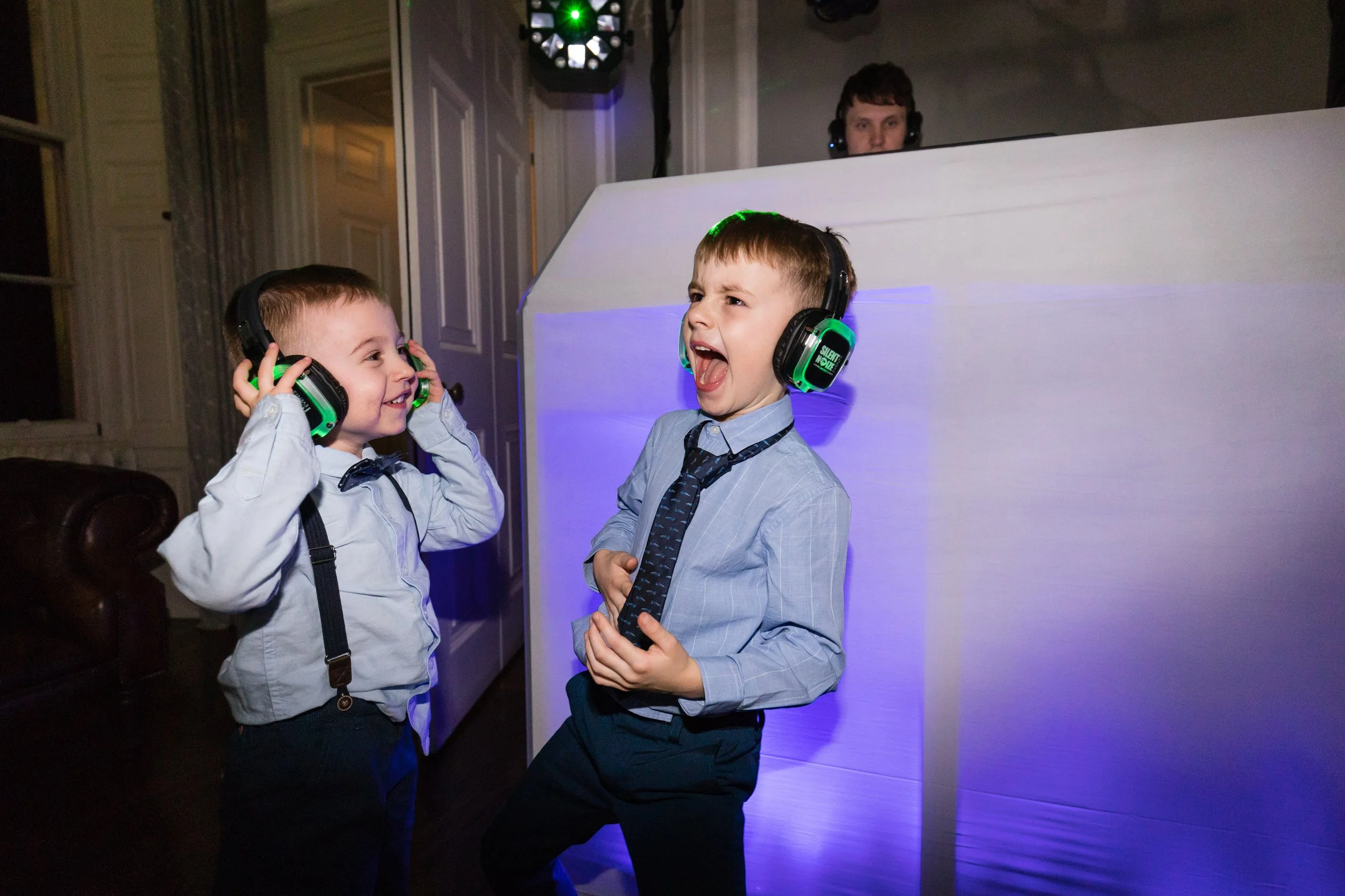 Two young boys at a party wearing headphones and dancing, with a DJ in the background.
