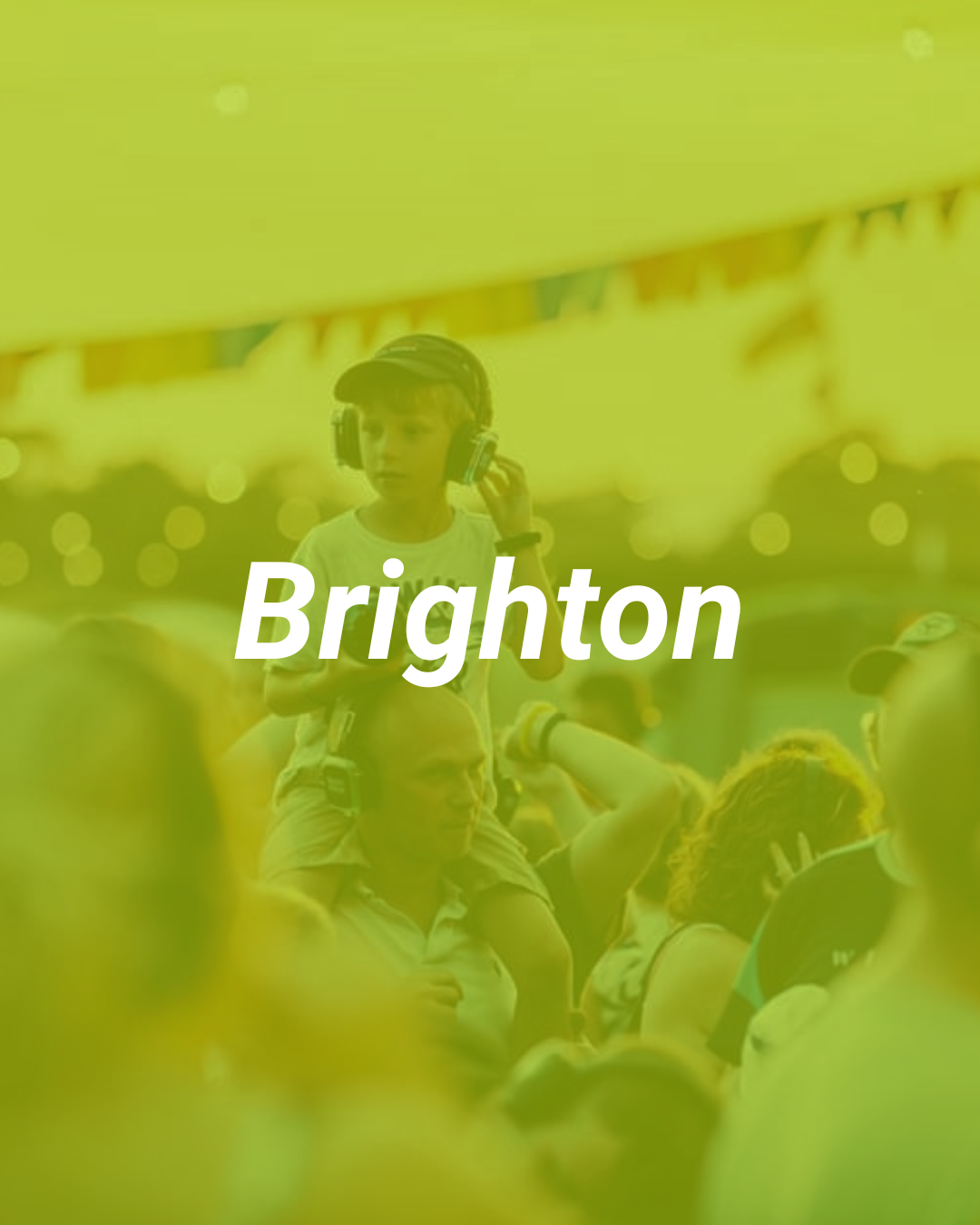 A young boy sitting on a man's shoulders, wearing headphones and a helmet at an outdoor event in Brighton, with other people around.