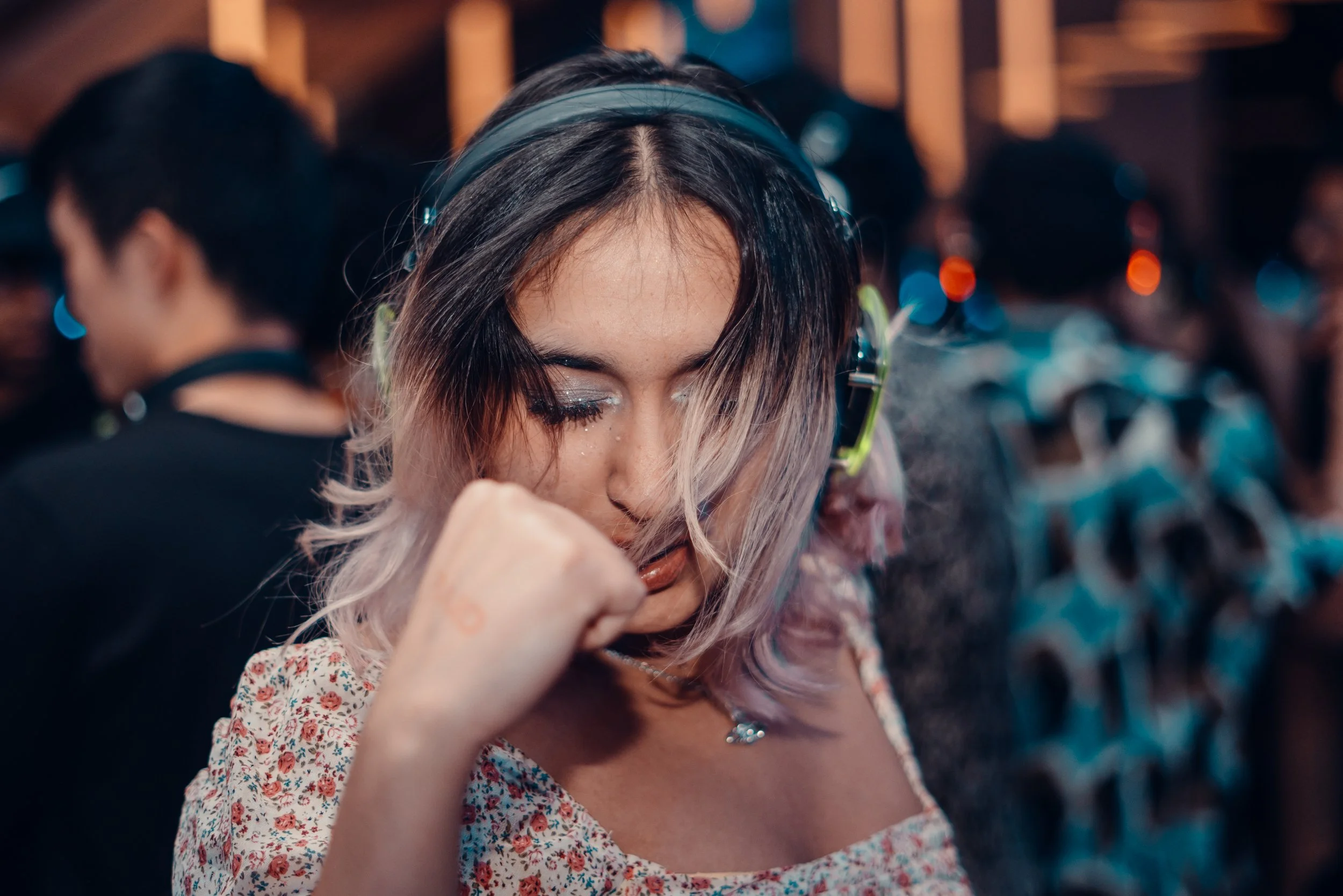 A young woman with light-colored hair, headphones, and sparkly makeup, appears to be lost in her music in a crowded indoor setting.