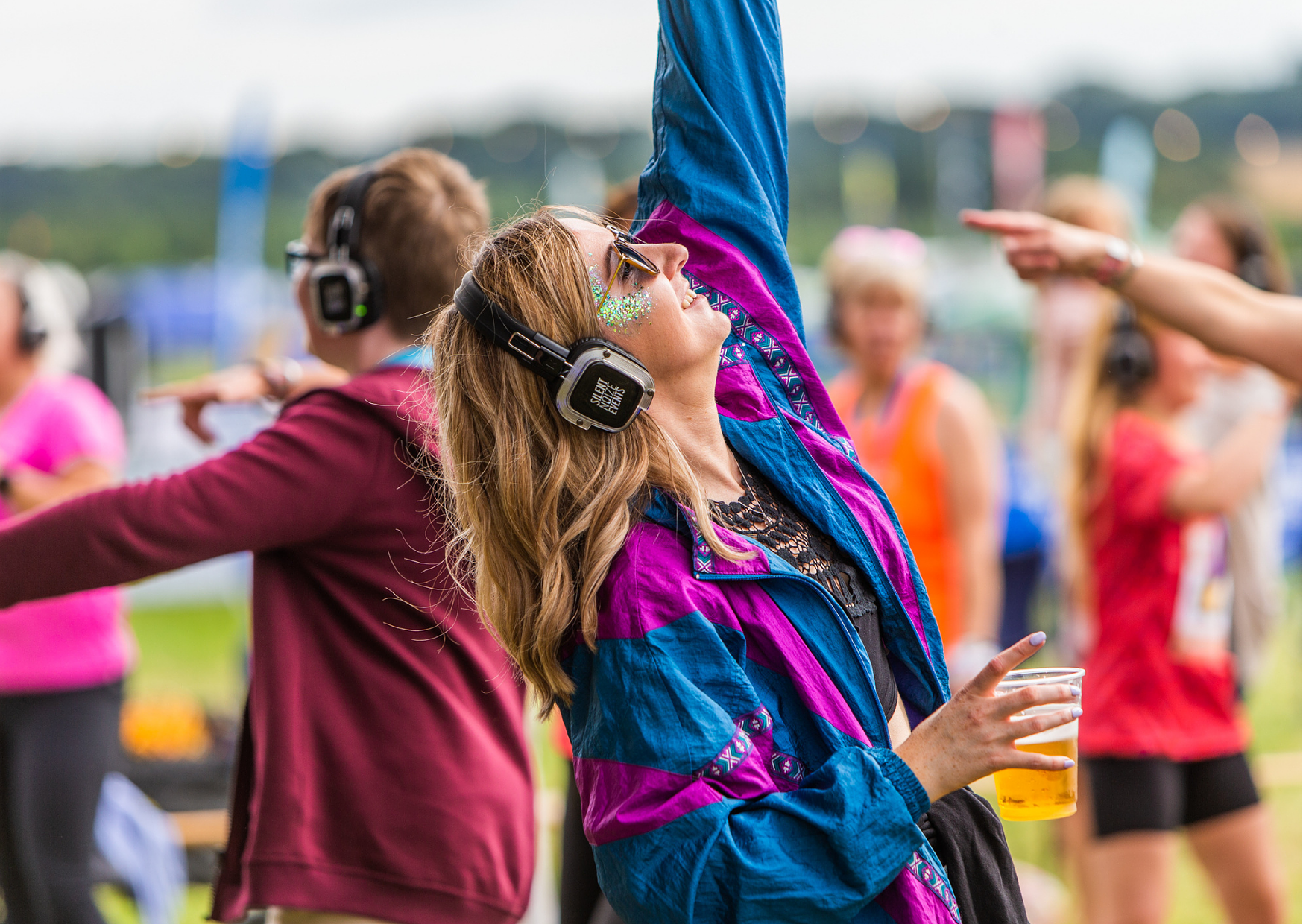 A woman at an outdoor event wearing headphones with 'Safety Center' written on them, holding a drink, and dancing with her eyes closed. She has glitter face paint and a colorful jacket, surrounded by other people also enjoying the event.