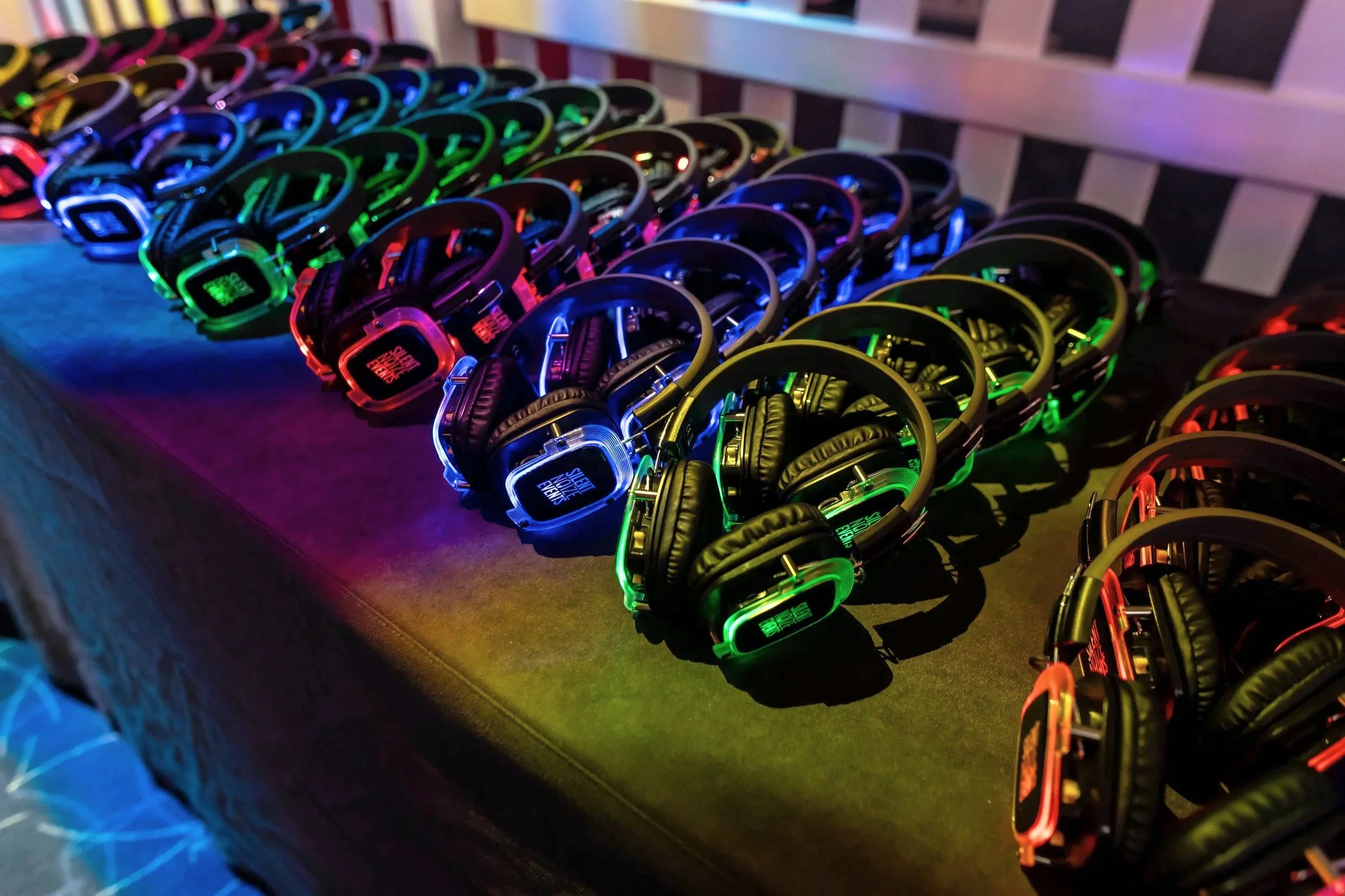 Multiple pairs of colorful glowing headphones arranged on a black table under multicolored lighting.