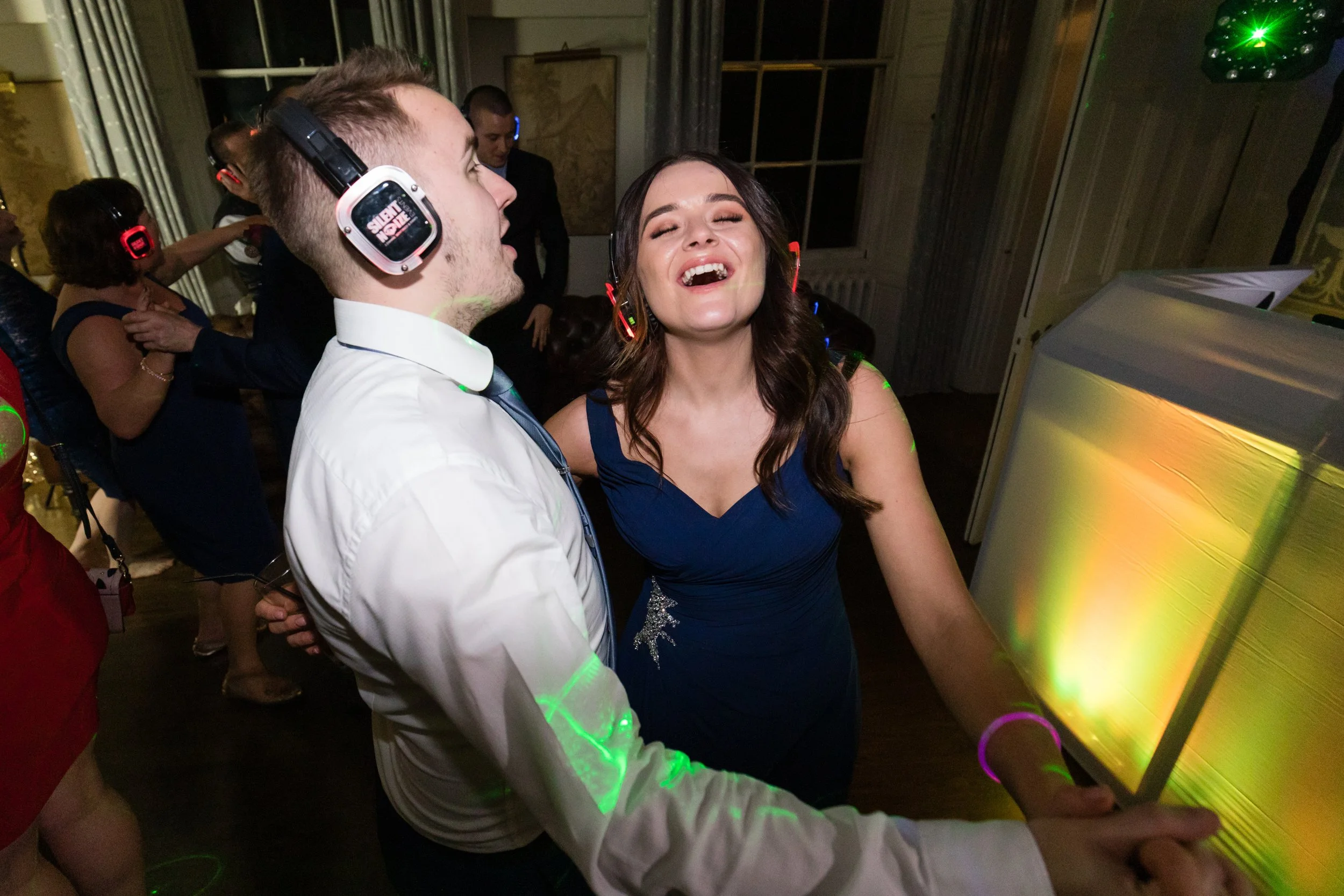 A group of people dancing and having fun at a party, with a woman in a blue dress singing along and a man in a white shirt wearing headphones, with colorful lighting and a DJ booth in the background.