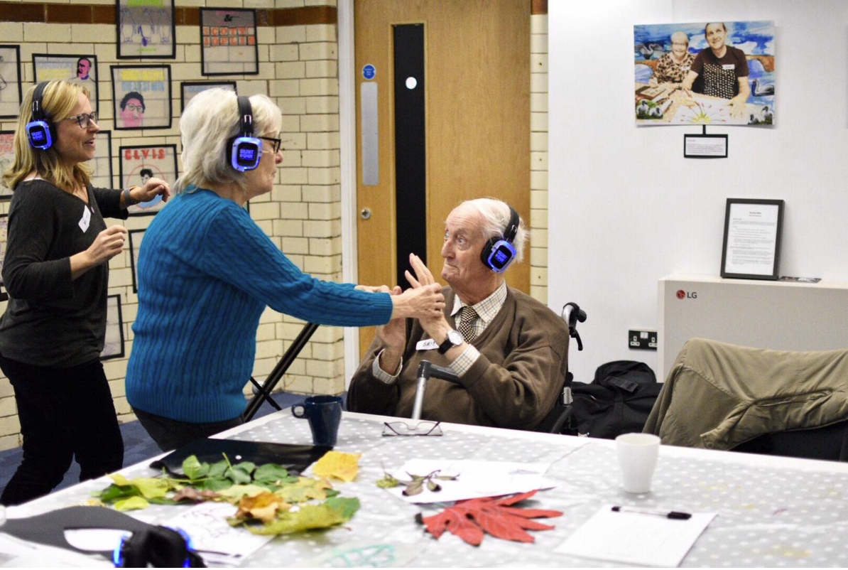 Two women and an elderly man with a walker, all wearing headphones, engaged in conversation in a room. The woman in blue is holding the man's hands, while another woman stands nearby. A table with leaves, papers, and cups is in the foreground, and walls adorned with framed pictures and artwork are in the background.