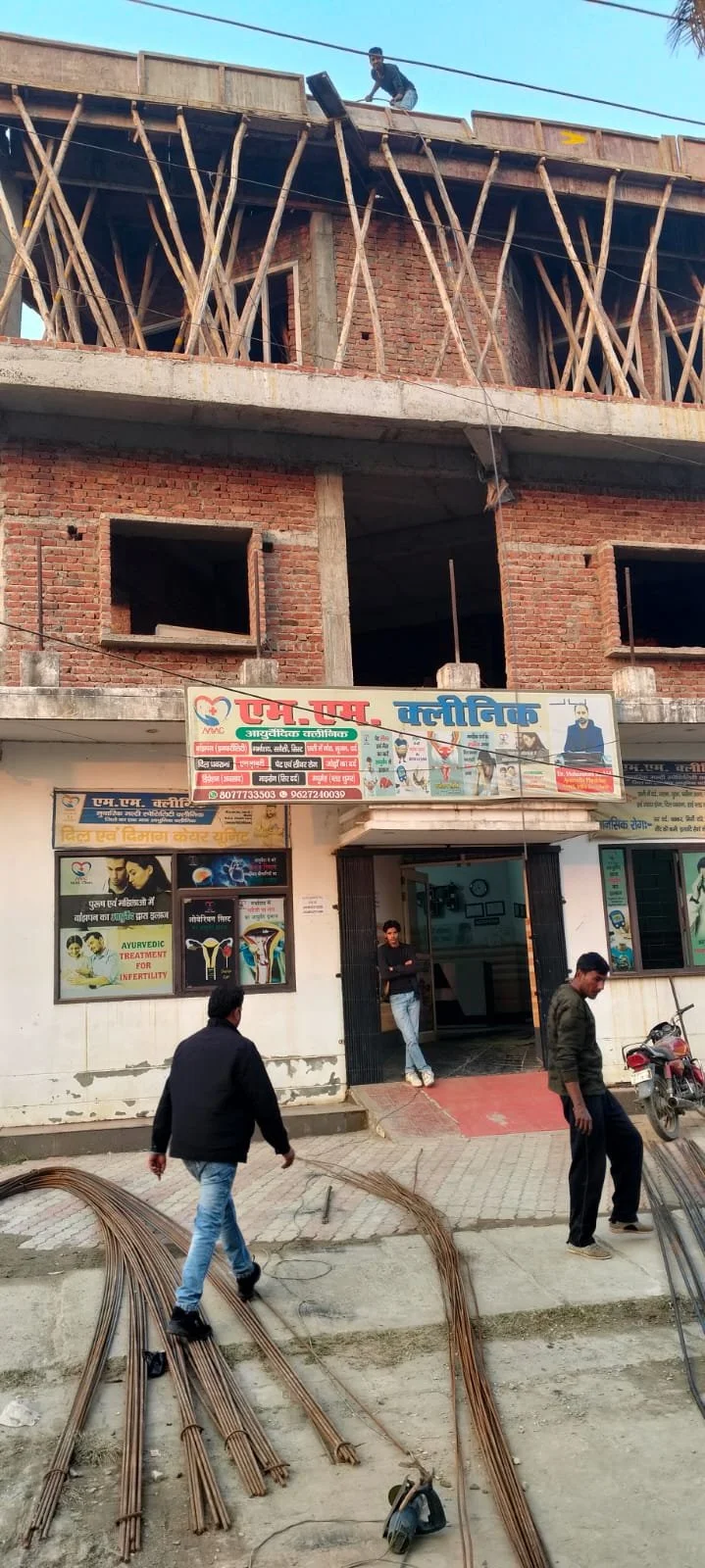 Construction site with unfinished brick building. Worker on the top floor working near the edge. Construction materials like steel rods on the ground. People walking past the site. Signboards in Hindi language on the building, indicating a medical or healthcare facility.