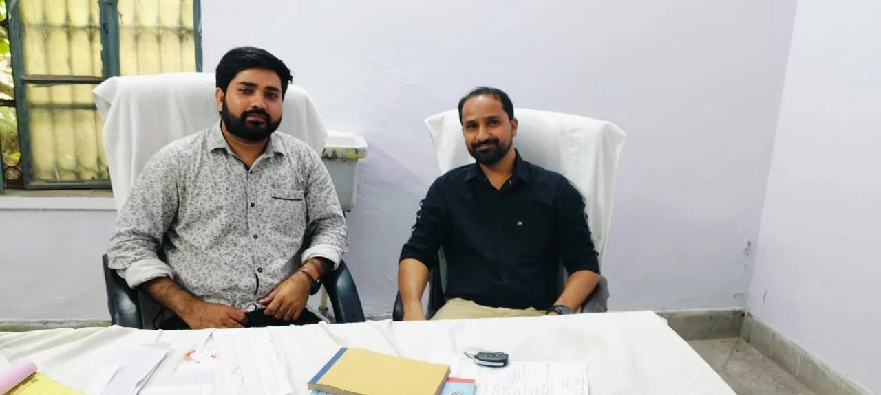 Two men sitting at a table in an office or meeting room, with documents and a mobile phone on the table, and a window in the background.