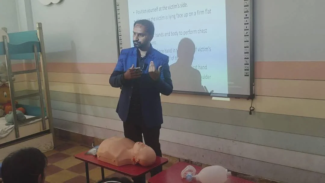 Man giving a CPR training session with a CPR dummy on a table, in front of a whiteboard with presentation slides. Classroom setting with lockers and stuffed toys in the background.