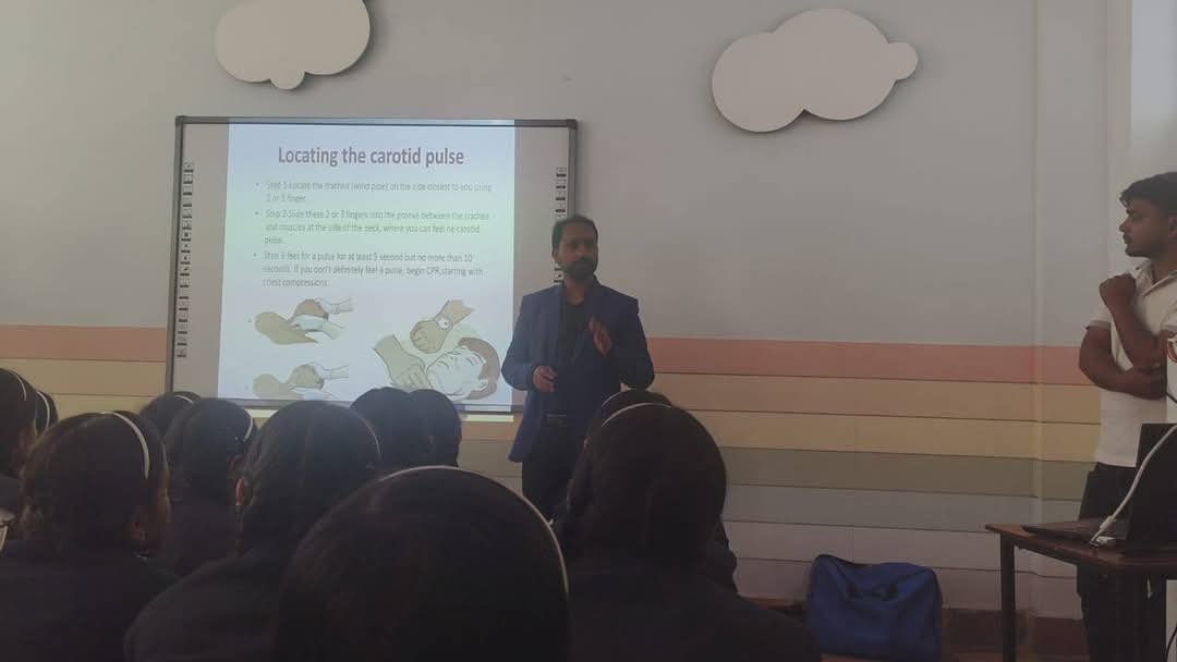 A teacher giving a presentation to students in a classroom, with a slide titled 'Locating the carotid pulse' visible on the screen.