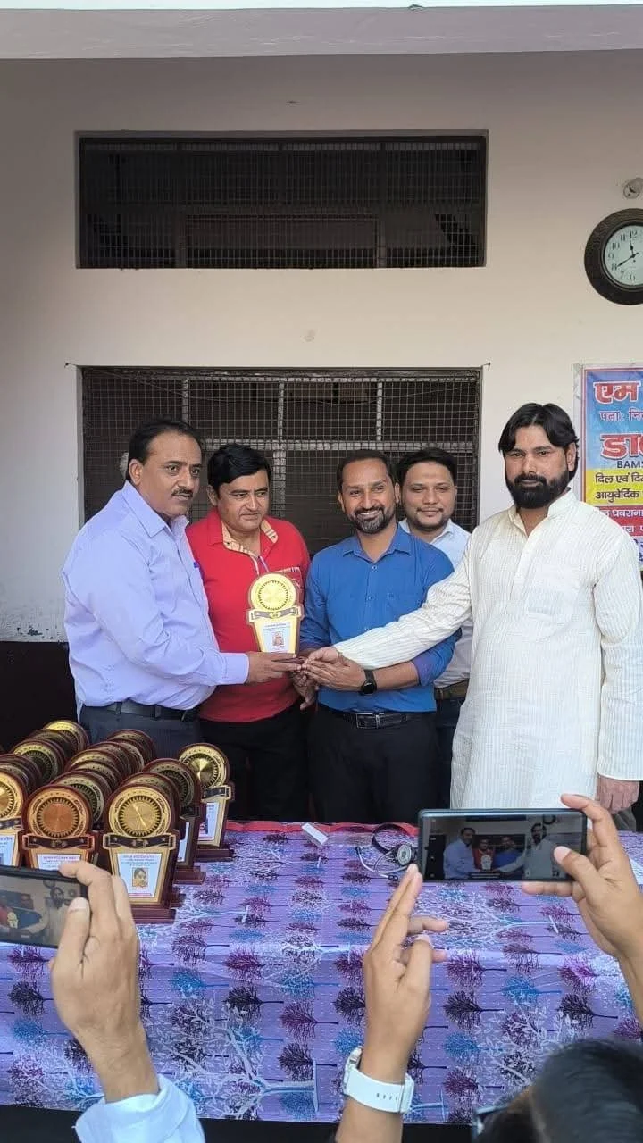 Group of five men at an award ceremony; one man is handing a trophy to another; trophies on the table in front; people taking photos; wall clock and posters in background.