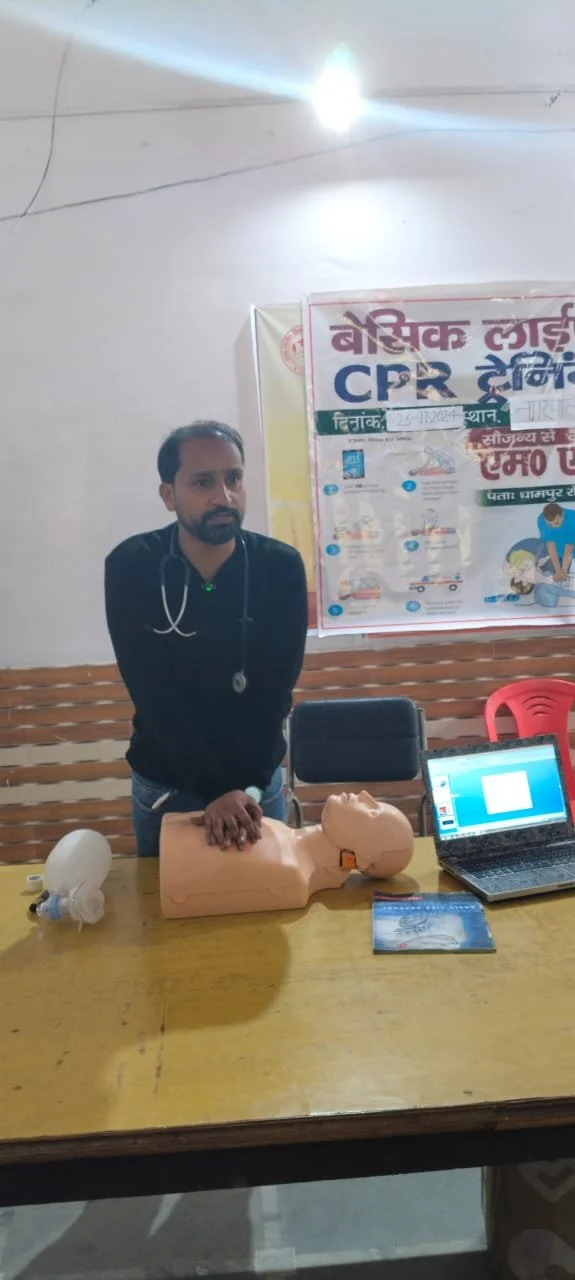 A man with a stethoscope demonstrating CPR on a mannequin in a classroom setting.