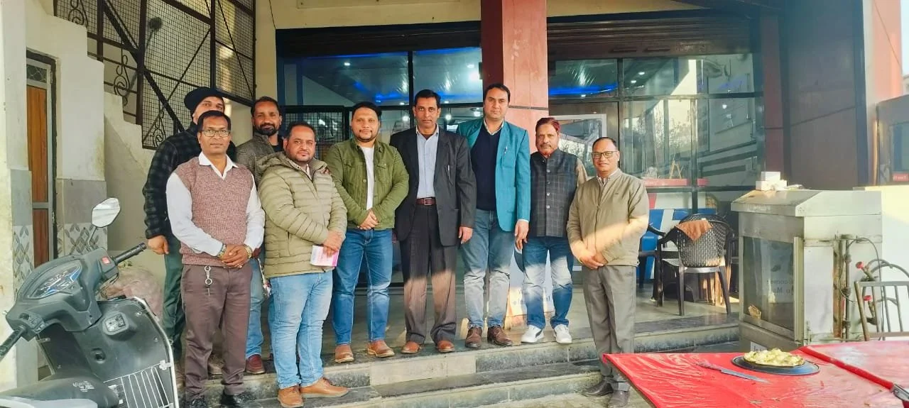 A group of eleven men standing together outside a building, with some holding papers, in casual and semi-formal attire. There is a scooter on the left and tables with chairs on the right, with a plate of food on a red tablecloth in the foreground.