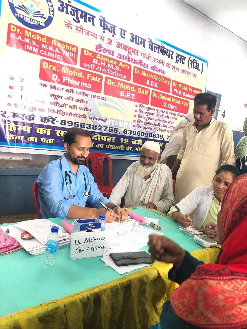 A doctor at a health camp signing documents while seated at a table with several people around him. There is a large banner in Hindi behind, listing medical professionals. The table has medical supplies, papers, and water bottles.