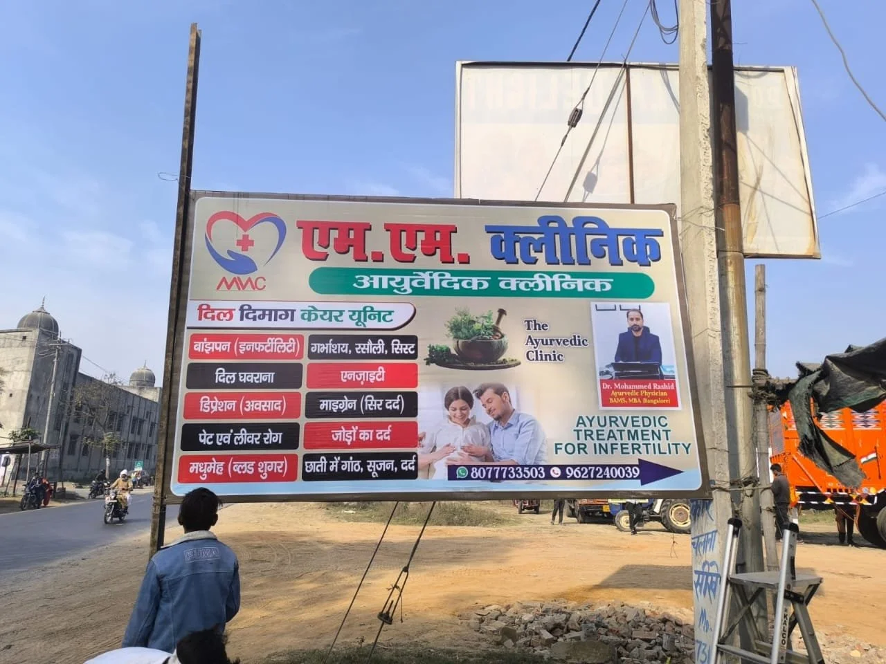 Billboard advertising an Ayurvedic clinic for infertility treatment, with images of a doctor, a couple, and various health benefits listed in Hindi, located outdoors near a road with people and vehicles.
