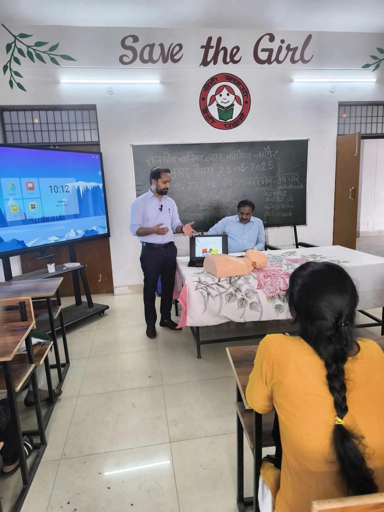 Teacher giving a demonstration to students in a classroom with a blackboard, screen, and 'Save the Girl' banner