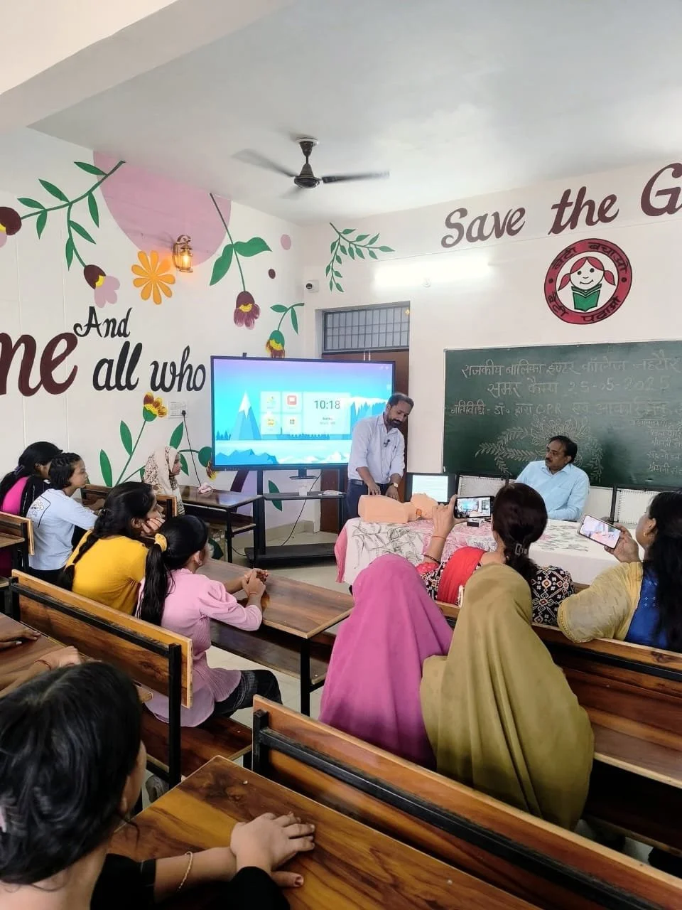 Classroom with students and teachers, decorated walls with floral murals, a blackboard, a large digital display, and people taking photos.