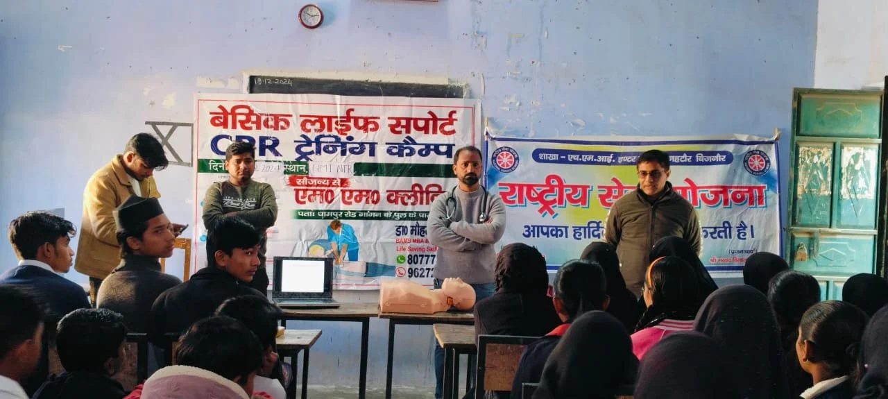 A classroom with a presentation on CPR training. Four men stand in front of a seated audience of students, with banners and a clock on the wall behind them.