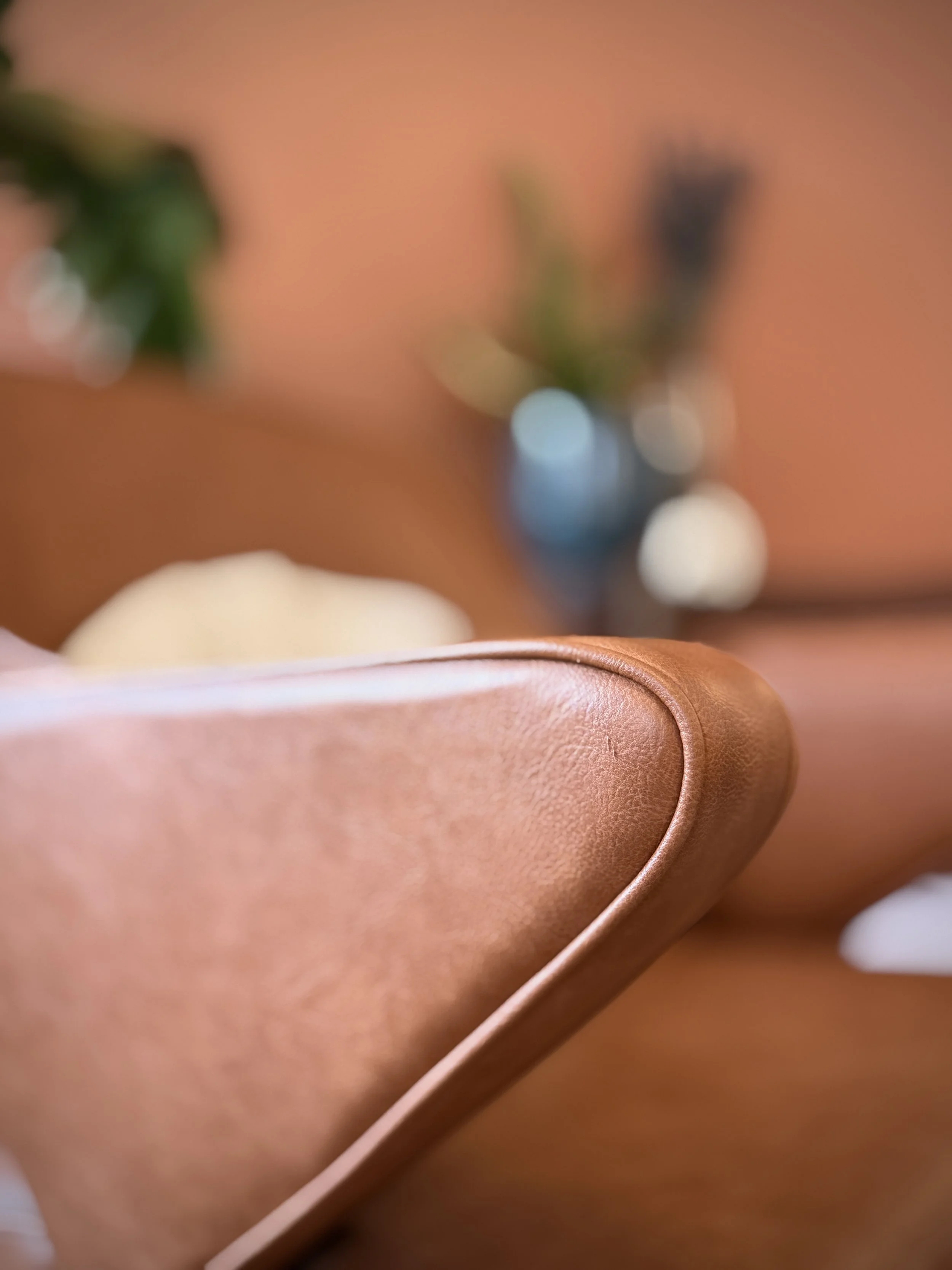 Close-up of a brown leather piece of furniture, likely a sofa or chair, with a blurred background featuring a blue vase with flowers and a soft pink wall.