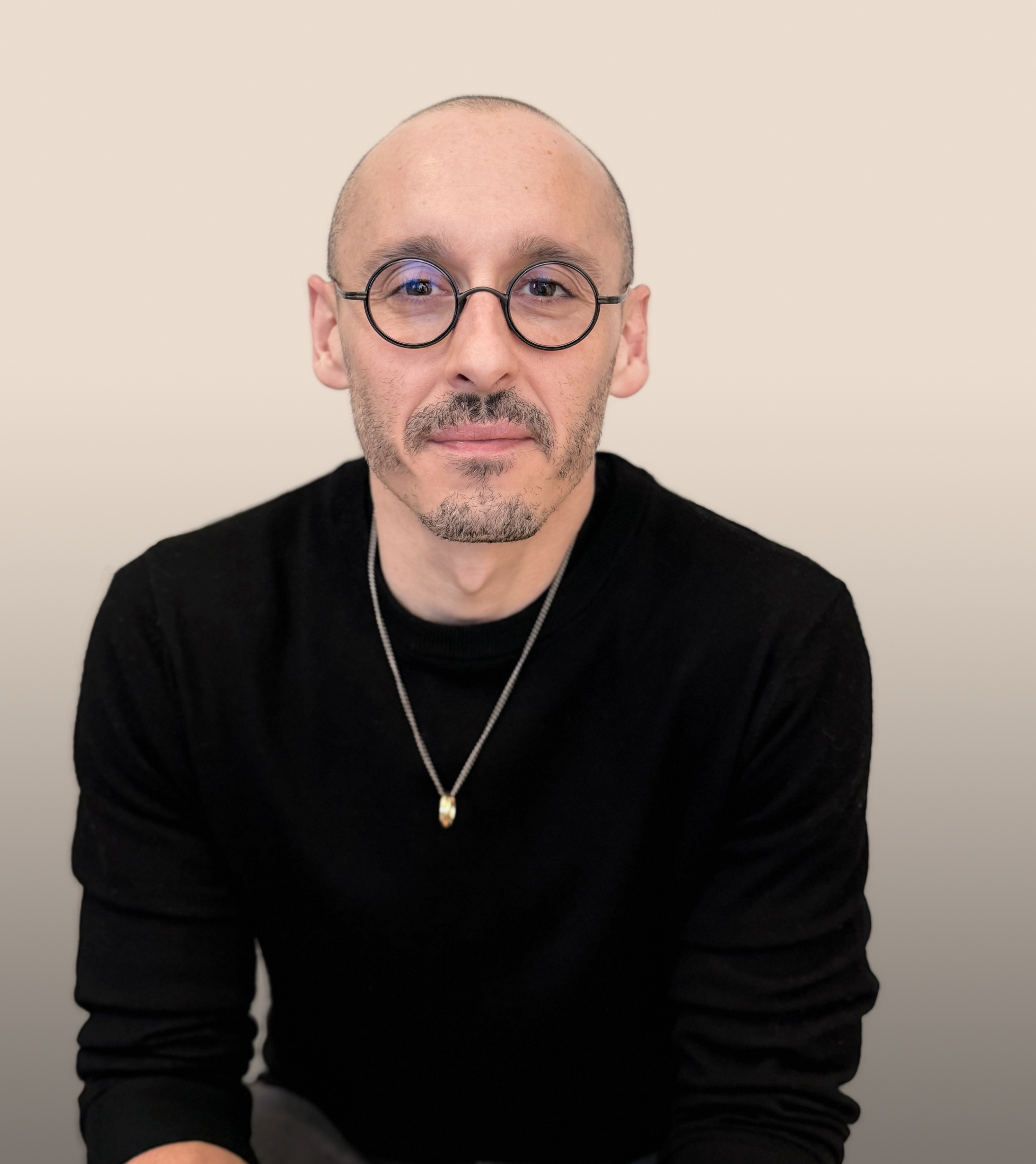 A man with glasses, a short beard, and a shaved head, wearing a black shirt and a necklace, posing against a white background.