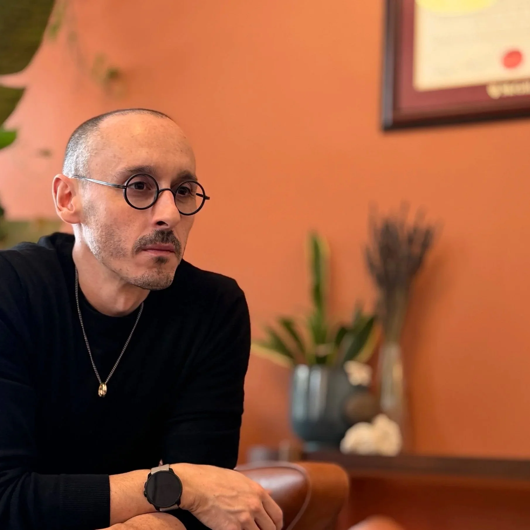 A man with glasses and a bald head, wearing a black shirt, is sitting indoors with an orange wall behind him. There is a framed certificate or picture on the wall and a vase with plants in the background.