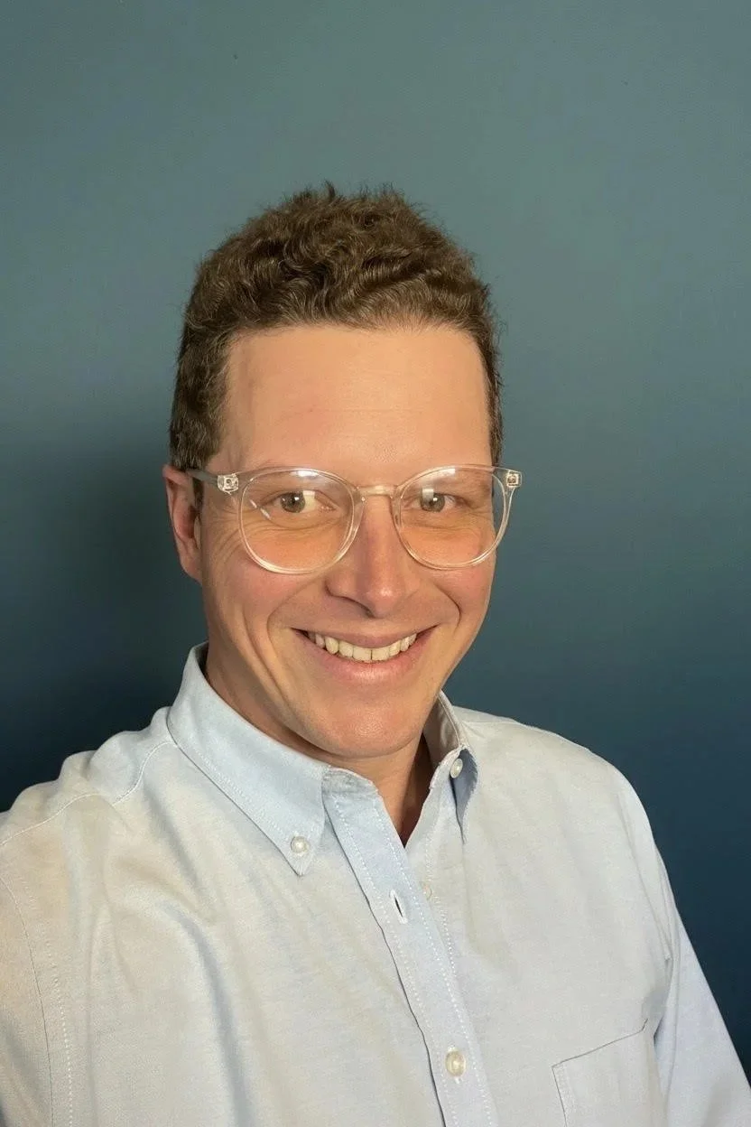 A man with short, curly brown hair wearing clear glasses and a light blue button-up shirt, smiling and standing against a blue background.