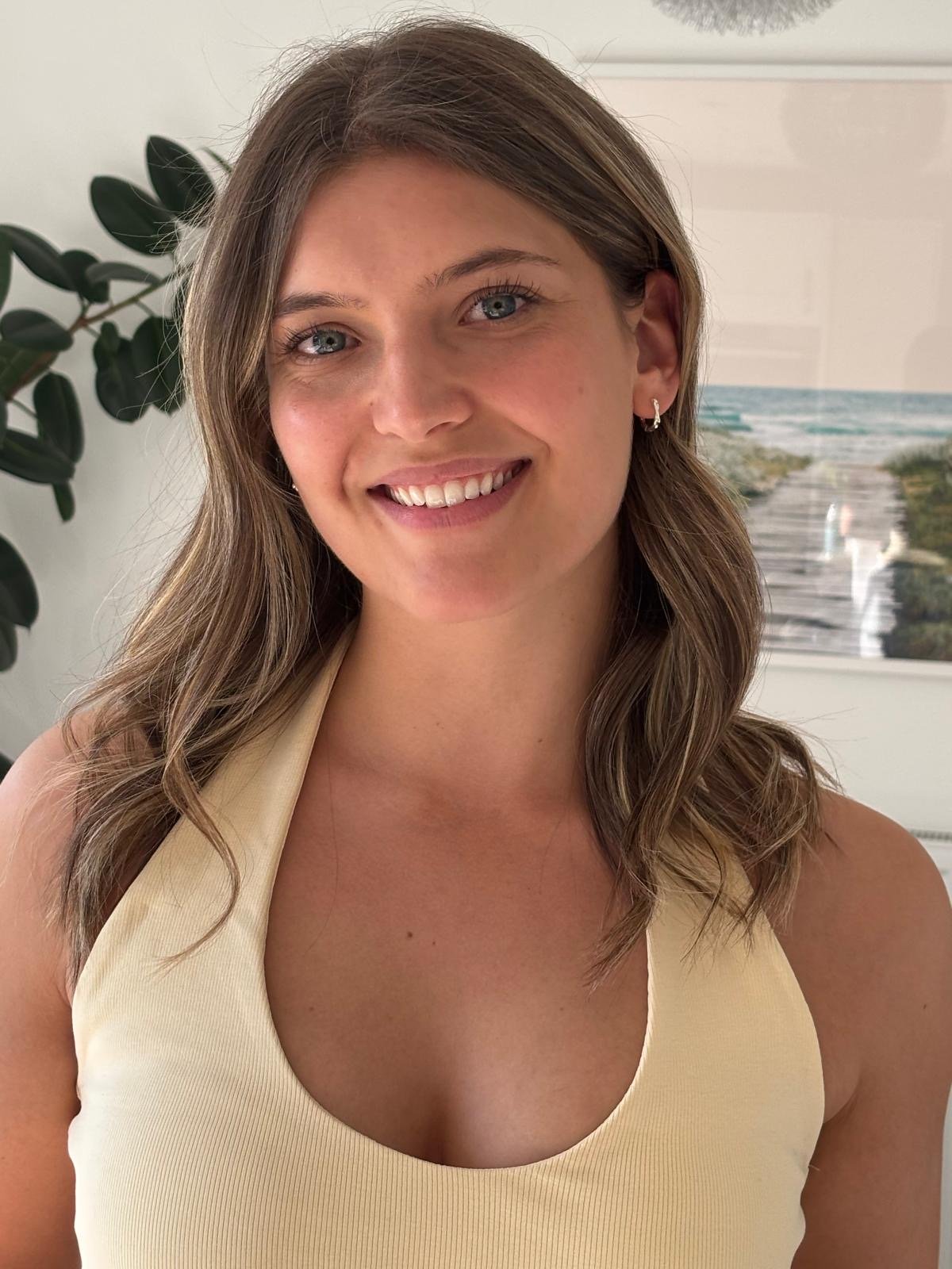 A smiling woman with long brown hair, blue eyes, and light skin, wearing a sleeveless yellow top, standing indoors with a large green plant and a beach scene picture in the background.
