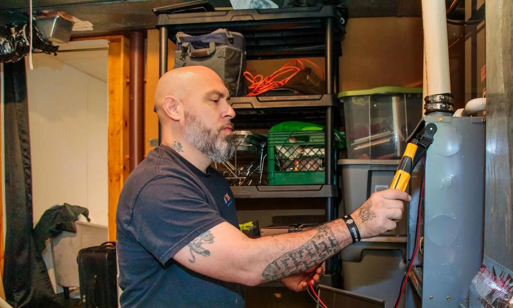 A man with tattoos on his arm and neck, bald head, and gray beard working on an electrical panel with a yellow screwdriver in a cluttered garage with tools and storage shelves.