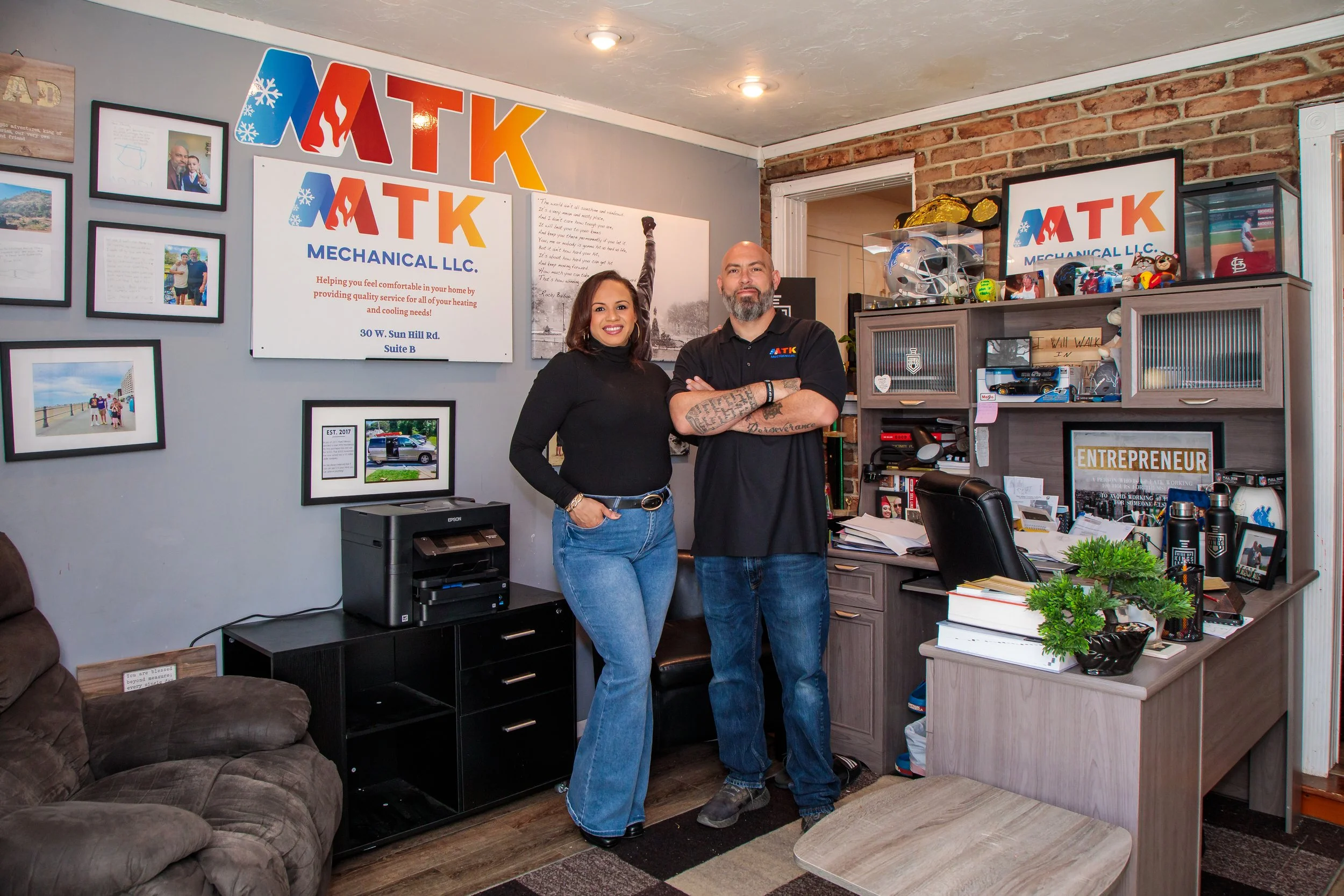 Two people standing in an office, smiling and posing for the camera. The woman on the left has shoulder-length hair and wears a black turtleneck and blue jeans. The man on the right has a beard, tattoos on his arms, and wears a black polo shirt and jeans. The office has a desk cluttered with papers, a shelf with various items, and walls decorated with framed photos, certificates, and a large sign that reads 'ATK MECHANICAL LLC.'