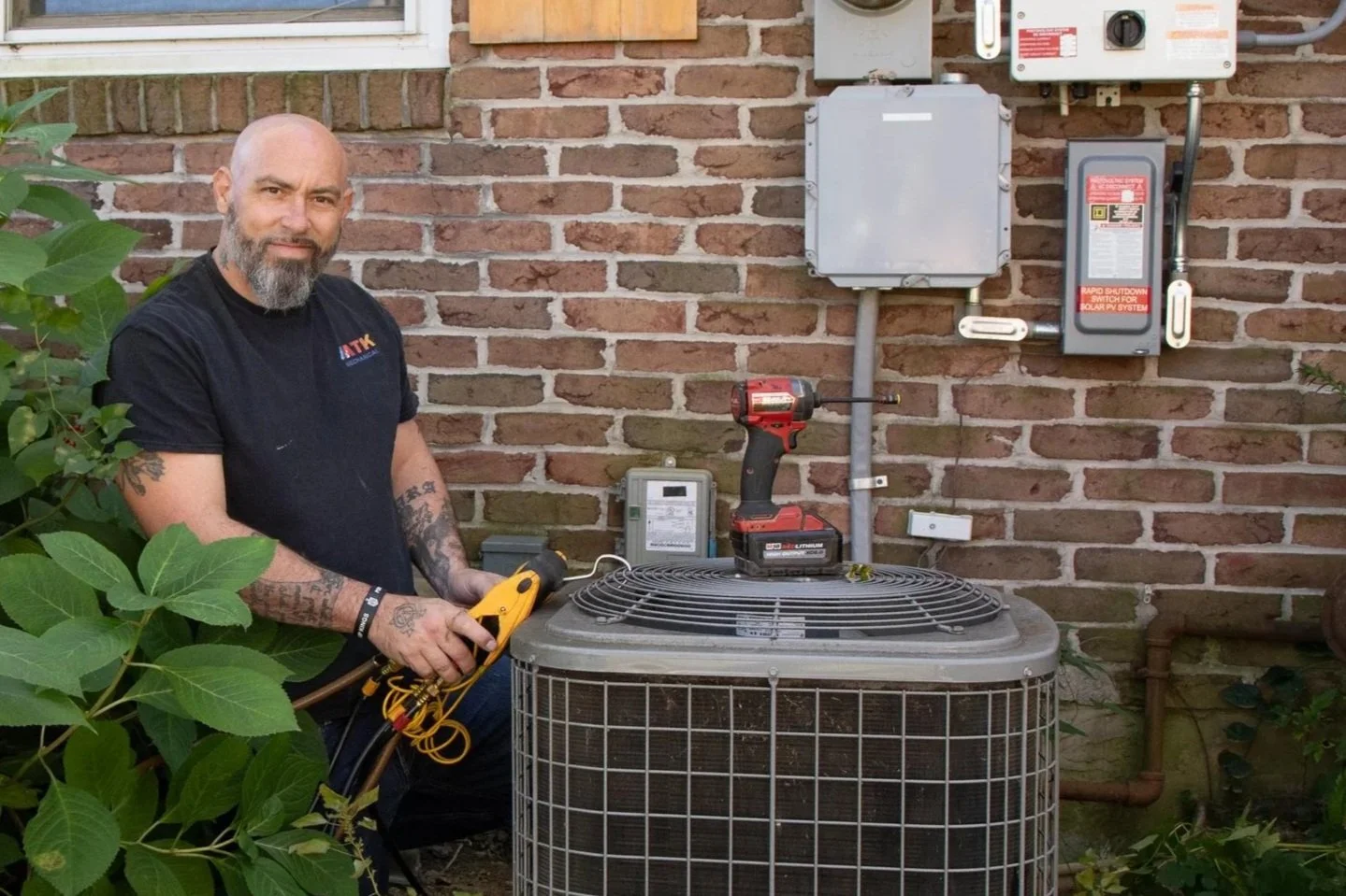 A man with a beard and tattooed arms, holding an HVAC multimeter, standing next to an outdoor air conditioning unit, with electrical panels and wires on a brick wall in the background.